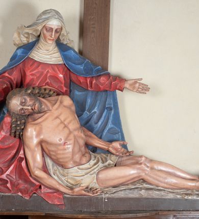 Zdjęcie nr 2: Rzeźba ustawiona na niewysokim, prostopadłościennym postumencie oraz na konsoli w kształcie fragmentu pilastra o kanelowanym trzonie i kapitelu dekorowanego motywem wolich oczu. Na tle krzyża z luźno przewieszoną przez poziomą belkę białą tkaniną ukazani Maria oraz martwy Chrystus. Jezus o szczupłym, muskularnym ciele z widocznymi śladami męki ukazany w pozycji półleżącej z torsem i ramionami wspartymi o postać Marii. Ręce ułożone luźno wzdłuż ciała. Głowa Chrystusa opada na prawe ramię postaci; twarz szczupła z wyraźnie zarysowanymi kośćmi policzkowymi, oczy zamknięte. Długie, brązowe włosy z przedziałkiem pośrodku głowy skręcają się w spiralne loki. Na twarzy krótki zarost. Wokół bioder Chrystusa luźno owinięte białe perizonium. Maria ukazana w pozycji klęczącej z rękoma uniesionymi na boki w dynamicznym geście. Głowa pochylona nieznacznie do dołu, w kierunku ciała Chrystusa. Twarz Marii szczupła, owalna z głęboko osadzonymi, dużymi oczami oraz wydatnym, prostym nosem. Postać ubrana jest w czerwoną suknię z długimi rękawami, niebieski płaszcz oraz białą chustę z podwiką wokół szyi. Po lewej stronie kompozycji, spod sukni widoczna jest bosa stopa.  