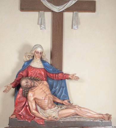 Zdjęcie nr 1: Rzeźba ustawiona na niewysokim, prostopadłościennym postumencie oraz na konsoli w kształcie fragmentu pilastra o kanelowanym trzonie i kapitelu dekorowanego motywem wolich oczu. Na tle krzyża z luźno przewieszoną przez poziomą belkę białą tkaniną ukazani Maria oraz martwy Chrystus. Jezus o szczupłym, muskularnym ciele z widocznymi śladami męki ukazany w pozycji półleżącej z torsem i ramionami wspartymi o postać Marii. Ręce ułożone luźno wzdłuż ciała. Głowa Chrystusa opada na prawe ramię postaci; twarz szczupła z wyraźnie zarysowanymi kośćmi policzkowymi, oczy zamknięte. Długie, brązowe włosy z przedziałkiem pośrodku głowy skręcają się w spiralne loki. Na twarzy krótki zarost. Wokół bioder Chrystusa luźno owinięte białe perizonium. Maria ukazana w pozycji klęczącej z rękoma uniesionymi na boki w dynamicznym geście. Głowa pochylona nieznacznie do dołu, w kierunku ciała Chrystusa. Twarz Marii szczupła, owalna z głęboko osadzonymi, dużymi oczami oraz wydatnym, prostym nosem. Postać ubrana jest w czerwoną suknię z długimi rękawami, niebieski płaszcz oraz białą chustę z podwiką wokół szyi. Po lewej stronie kompozycji, spod sukni widoczna jest bosa stopa.  