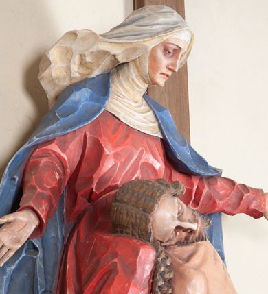 Zdjęcie nr 7: Rzeźba ustawiona na niewysokim, prostopadłościennym postumencie oraz na konsoli w kształcie fragmentu pilastra o kanelowanym trzonie i kapitelu dekorowanego motywem wolich oczu. Na tle krzyża z luźno przewieszoną przez poziomą belkę białą tkaniną ukazani Maria oraz martwy Chrystus. Jezus o szczupłym, muskularnym ciele z widocznymi śladami męki ukazany w pozycji półleżącej z torsem i ramionami wspartymi o postać Marii. Ręce ułożone luźno wzdłuż ciała. Głowa Chrystusa opada na prawe ramię postaci; twarz szczupła z wyraźnie zarysowanymi kośćmi policzkowymi, oczy zamknięte. Długie, brązowe włosy z przedziałkiem pośrodku głowy skręcają się w spiralne loki. Na twarzy krótki zarost. Wokół bioder Chrystusa luźno owinięte białe perizonium. Maria ukazana w pozycji klęczącej z rękoma uniesionymi na boki w dynamicznym geście. Głowa pochylona nieznacznie do dołu, w kierunku ciała Chrystusa. Twarz Marii szczupła, owalna z głęboko osadzonymi, dużymi oczami oraz wydatnym, prostym nosem. Postać ubrana jest w czerwoną suknię z długimi rękawami, niebieski płaszcz oraz białą chustę z podwiką wokół szyi. Po lewej stronie kompozycji, spod sukni widoczna jest bosa stopa.  