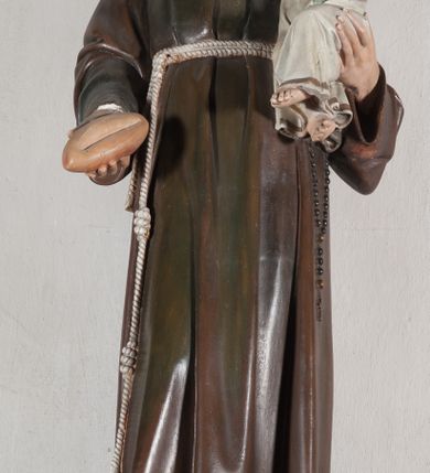 Zdjęcie nr 11: Rzeźba pełnoplastyczna, umieszczona na niewielkim prostopadłościennym postumencie o ściętych narożach oraz złoconej i srebrzonej konsoli utworzonej z liści akantu i róż. Święty ukazany całopostaciowo, frontalnie w pozycji stojącej. Antoni został przedstawiony jako młody mężczyzna o owalnej, pozbawionej zarostu twarzy i krótkich, brązowych włosach z tonsurą. Ma oczy wpatrzone przed siebie, wydatny, prosty nos, usta zamknięte. Postać w prawej ręce trzyma bochenek chleba, a na lewej podtrzymuje Dzieciątko Jezus. Święty ubrany jest w długi, ciemnobrązowy habit franciszkański. Do sznura z trzema węzłami ujmującego go w pasie przywiązany jest różaniec. Spod habitu widoczne stopy obute w sandały. U stóp ustawiony kosz z bochenkami chleba. Jasnowłose Dzieciątko Jezus ukazane w pozycji siedzącej z głową zwróconą w trzech czwartych w prawo. Prawą ręką wykonuje gest błogosławieństwa, lewą składa na piersi, podtrzymując kwiat białej lilii. Postać ubrana z białą tunikę ze złotą lamówką wokół dekoltu, rękawów oraz dolnej krawędzi szaty. Na konsoli, na której ustawiona jest rzeźba widnieje napis: ŚWIĘTY ANTONI MÓDL SIĘ ZA NAMI.