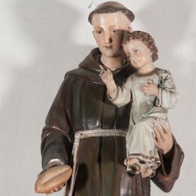 Zdjęcie nr 1: Rzeźba pełnoplastyczna, umieszczona na niewielkim prostopadłościennym postumencie o ściętych narożach oraz złoconej i srebrzonej konsoli utworzonej z liści akantu i róż. Święty ukazany całopostaciowo, frontalnie w pozycji stojącej. Antoni został przedstawiony jako młody mężczyzna o owalnej, pozbawionej zarostu twarzy i krótkich, brązowych włosach z tonsurą. Ma oczy wpatrzone przed siebie, wydatny, prosty nos, usta zamknięte. Postać w prawej ręce trzyma bochenek chleba, a na lewej podtrzymuje Dzieciątko Jezus. Święty ubrany jest w długi, ciemnobrązowy habit franciszkański. Do sznura z trzema węzłami ujmującego go w pasie przywiązany jest różaniec. Spod habitu widoczne stopy obute w sandały. U stóp ustawiony kosz z bochenkami chleba. Jasnowłose Dzieciątko Jezus ukazane w pozycji siedzącej z głową zwróconą w trzech czwartych w prawo. Prawą ręką wykonuje gest błogosławieństwa, lewą składa na piersi, podtrzymując kwiat białej lilii. Postać ubrana z białą tunikę ze złotą lamówką wokół dekoltu, rękawów oraz dolnej krawędzi szaty. Na konsoli, na której ustawiona jest rzeźba widnieje napis: ŚWIĘTY ANTONI MÓDL SIĘ ZA NAMI.