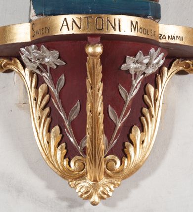 Zdjęcie nr 8: Rzeźba pełnoplastyczna, umieszczona na niewielkim prostopadłościennym postumencie o ściętych narożach oraz złoconej i srebrzonej konsoli utworzonej z liści akantu i róż. Święty ukazany całopostaciowo, frontalnie w pozycji stojącej. Antoni został przedstawiony jako młody mężczyzna o owalnej, pozbawionej zarostu twarzy i krótkich, brązowych włosach z tonsurą. Ma oczy wpatrzone przed siebie, wydatny, prosty nos, usta zamknięte. Postać w prawej ręce trzyma bochenek chleba, a na lewej podtrzymuje Dzieciątko Jezus. Święty ubrany jest w długi, ciemnobrązowy habit franciszkański. Do sznura z trzema węzłami ujmującego go w pasie przywiązany jest różaniec. Spod habitu widoczne stopy obute w sandały. U stóp ustawiony kosz z bochenkami chleba. Jasnowłose Dzieciątko Jezus ukazane w pozycji siedzącej z głową zwróconą w trzech czwartych w prawo. Prawą ręką wykonuje gest błogosławieństwa, lewą składa na piersi, podtrzymując kwiat białej lilii. Postać ubrana z białą tunikę ze złotą lamówką wokół dekoltu, rękawów oraz dolnej krawędzi szaty. Na konsoli, na której ustawiona jest rzeźba widnieje napis: ŚWIĘTY ANTONI MÓDL SIĘ ZA NAMI.