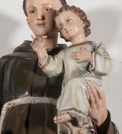 Zdjęcie nr 3: Rzeźba pełnoplastyczna, umieszczona na niewielkim prostopadłościennym postumencie o ściętych narożach oraz złoconej i srebrzonej konsoli utworzonej z liści akantu i róż. Święty ukazany całopostaciowo, frontalnie w pozycji stojącej. Antoni został przedstawiony jako młody mężczyzna o owalnej, pozbawionej zarostu twarzy i krótkich, brązowych włosach z tonsurą. Ma oczy wpatrzone przed siebie, wydatny, prosty nos, usta zamknięte. Postać w prawej ręce trzyma bochenek chleba, a na lewej podtrzymuje Dzieciątko Jezus. Święty ubrany jest w długi, ciemnobrązowy habit franciszkański. Do sznura z trzema węzłami ujmującego go w pasie przywiązany jest różaniec. Spod habitu widoczne stopy obute w sandały. U stóp ustawiony kosz z bochenkami chleba. Jasnowłose Dzieciątko Jezus ukazane w pozycji siedzącej z głową zwróconą w trzech czwartych w prawo. Prawą ręką wykonuje gest błogosławieństwa, lewą składa na piersi, podtrzymując kwiat białej lilii. Postać ubrana z białą tunikę ze złotą lamówką wokół dekoltu, rękawów oraz dolnej krawędzi szaty. Na konsoli, na której ustawiona jest rzeźba widnieje napis: ŚWIĘTY ANTONI MÓDL SIĘ ZA NAMI.