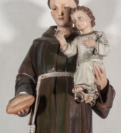 Zdjęcie nr 16: Rzeźba pełnoplastyczna, umieszczona na niewielkim prostopadłościennym postumencie o ściętych narożach oraz złoconej i srebrzonej konsoli utworzonej z liści akantu i róż. Święty ukazany całopostaciowo, frontalnie w pozycji stojącej. Antoni został przedstawiony jako młody mężczyzna o owalnej, pozbawionej zarostu twarzy i krótkich, brązowych włosach z tonsurą. Ma oczy wpatrzone przed siebie, wydatny, prosty nos, usta zamknięte. Postać w prawej ręce trzyma bochenek chleba, a na lewej podtrzymuje Dzieciątko Jezus. Święty ubrany jest w długi, ciemnobrązowy habit franciszkański. Do sznura z trzema węzłami ujmującego go w pasie przywiązany jest różaniec. Spod habitu widoczne stopy obute w sandały. U stóp ustawiony kosz z bochenkami chleba. Jasnowłose Dzieciątko Jezus ukazane w pozycji siedzącej z głową zwróconą w trzech czwartych w prawo. Prawą ręką wykonuje gest błogosławieństwa, lewą składa na piersi, podtrzymując kwiat białej lilii. Postać ubrana z białą tunikę ze złotą lamówką wokół dekoltu, rękawów oraz dolnej krawędzi szaty. Na konsoli, na której ustawiona jest rzeźba widnieje napis: ŚWIĘTY ANTONI MÓDL SIĘ ZA NAMI.