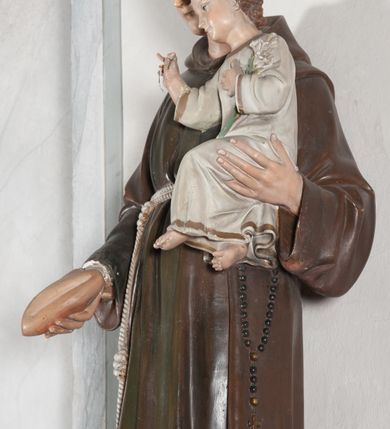 Zdjęcie nr 14: Rzeźba pełnoplastyczna, umieszczona na niewielkim prostopadłościennym postumencie o ściętych narożach oraz złoconej i srebrzonej konsoli utworzonej z liści akantu i róż. Święty ukazany całopostaciowo, frontalnie w pozycji stojącej. Antoni został przedstawiony jako młody mężczyzna o owalnej, pozbawionej zarostu twarzy i krótkich, brązowych włosach z tonsurą. Ma oczy wpatrzone przed siebie, wydatny, prosty nos, usta zamknięte. Postać w prawej ręce trzyma bochenek chleba, a na lewej podtrzymuje Dzieciątko Jezus. Święty ubrany jest w długi, ciemnobrązowy habit franciszkański. Do sznura z trzema węzłami ujmującego go w pasie przywiązany jest różaniec. Spod habitu widoczne stopy obute w sandały. U stóp ustawiony kosz z bochenkami chleba. Jasnowłose Dzieciątko Jezus ukazane w pozycji siedzącej z głową zwróconą w trzech czwartych w prawo. Prawą ręką wykonuje gest błogosławieństwa, lewą składa na piersi, podtrzymując kwiat białej lilii. Postać ubrana z białą tunikę ze złotą lamówką wokół dekoltu, rękawów oraz dolnej krawędzi szaty. Na konsoli, na której ustawiona jest rzeźba widnieje napis: ŚWIĘTY ANTONI MÓDL SIĘ ZA NAMI.