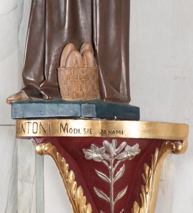 Zdjęcie nr 6: Rzeźba pełnoplastyczna, umieszczona na niewielkim prostopadłościennym postumencie o ściętych narożach oraz złoconej i srebrzonej konsoli utworzonej z liści akantu i róż. Święty ukazany całopostaciowo, frontalnie w pozycji stojącej. Antoni został przedstawiony jako młody mężczyzna o owalnej, pozbawionej zarostu twarzy i krótkich, brązowych włosach z tonsurą. Ma oczy wpatrzone przed siebie, wydatny, prosty nos, usta zamknięte. Postać w prawej ręce trzyma bochenek chleba, a na lewej podtrzymuje Dzieciątko Jezus. Święty ubrany jest w długi, ciemnobrązowy habit franciszkański. Do sznura z trzema węzłami ujmującego go w pasie przywiązany jest różaniec. Spod habitu widoczne stopy obute w sandały. U stóp ustawiony kosz z bochenkami chleba. Jasnowłose Dzieciątko Jezus ukazane w pozycji siedzącej z głową zwróconą w trzech czwartych w prawo. Prawą ręką wykonuje gest błogosławieństwa, lewą składa na piersi, podtrzymując kwiat białej lilii. Postać ubrana z białą tunikę ze złotą lamówką wokół dekoltu, rękawów oraz dolnej krawędzi szaty. Na konsoli, na której ustawiona jest rzeźba widnieje napis: ŚWIĘTY ANTONI MÓDL SIĘ ZA NAMI.