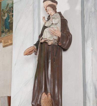 Zdjęcie nr 9: Rzeźba pełnoplastyczna, umieszczona na niewielkim prostopadłościennym postumencie o ściętych narożach oraz złoconej i srebrzonej konsoli utworzonej z liści akantu i róż. Święty ukazany całopostaciowo, frontalnie w pozycji stojącej. Antoni został przedstawiony jako młody mężczyzna o owalnej, pozbawionej zarostu twarzy i krótkich, brązowych włosach z tonsurą. Ma oczy wpatrzone przed siebie, wydatny, prosty nos, usta zamknięte. Postać w prawej ręce trzyma bochenek chleba, a na lewej podtrzymuje Dzieciątko Jezus. Święty ubrany jest w długi, ciemnobrązowy habit franciszkański. Do sznura z trzema węzłami ujmującego go w pasie przywiązany jest różaniec. Spod habitu widoczne stopy obute w sandały. U stóp ustawiony kosz z bochenkami chleba. Jasnowłose Dzieciątko Jezus ukazane w pozycji siedzącej z głową zwróconą w trzech czwartych w prawo. Prawą ręką wykonuje gest błogosławieństwa, lewą składa na piersi, podtrzymując kwiat białej lilii. Postać ubrana z białą tunikę ze złotą lamówką wokół dekoltu, rękawów oraz dolnej krawędzi szaty. Na konsoli, na której ustawiona jest rzeźba widnieje napis: ŚWIĘTY ANTONI MÓDL SIĘ ZA NAMI.