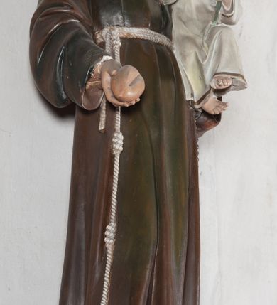 Zdjęcie nr 12: Rzeźba pełnoplastyczna, umieszczona na niewielkim prostopadłościennym postumencie o ściętych narożach oraz złoconej i srebrzonej konsoli utworzonej z liści akantu i róż. Święty ukazany całopostaciowo, frontalnie w pozycji stojącej. Antoni został przedstawiony jako młody mężczyzna o owalnej, pozbawionej zarostu twarzy i krótkich, brązowych włosach z tonsurą. Ma oczy wpatrzone przed siebie, wydatny, prosty nos, usta zamknięte. Postać w prawej ręce trzyma bochenek chleba, a na lewej podtrzymuje Dzieciątko Jezus. Święty ubrany jest w długi, ciemnobrązowy habit franciszkański. Do sznura z trzema węzłami ujmującego go w pasie przywiązany jest różaniec. Spod habitu widoczne stopy obute w sandały. U stóp ustawiony kosz z bochenkami chleba. Jasnowłose Dzieciątko Jezus ukazane w pozycji siedzącej z głową zwróconą w trzech czwartych w prawo. Prawą ręką wykonuje gest błogosławieństwa, lewą składa na piersi, podtrzymując kwiat białej lilii. Postać ubrana z białą tunikę ze złotą lamówką wokół dekoltu, rękawów oraz dolnej krawędzi szaty. Na konsoli, na której ustawiona jest rzeźba widnieje napis: ŚWIĘTY ANTONI MÓDL SIĘ ZA NAMI.