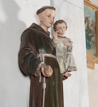 Zdjęcie nr 13: Rzeźba pełnoplastyczna, umieszczona na niewielkim prostopadłościennym postumencie o ściętych narożach oraz złoconej i srebrzonej konsoli utworzonej z liści akantu i róż. Święty ukazany całopostaciowo, frontalnie w pozycji stojącej. Antoni został przedstawiony jako młody mężczyzna o owalnej, pozbawionej zarostu twarzy i krótkich, brązowych włosach z tonsurą. Ma oczy wpatrzone przed siebie, wydatny, prosty nos, usta zamknięte. Postać w prawej ręce trzyma bochenek chleba, a na lewej podtrzymuje Dzieciątko Jezus. Święty ubrany jest w długi, ciemnobrązowy habit franciszkański. Do sznura z trzema węzłami ujmującego go w pasie przywiązany jest różaniec. Spod habitu widoczne stopy obute w sandały. U stóp ustawiony kosz z bochenkami chleba. Jasnowłose Dzieciątko Jezus ukazane w pozycji siedzącej z głową zwróconą w trzech czwartych w prawo. Prawą ręką wykonuje gest błogosławieństwa, lewą składa na piersi, podtrzymując kwiat białej lilii. Postać ubrana z białą tunikę ze złotą lamówką wokół dekoltu, rękawów oraz dolnej krawędzi szaty. Na konsoli, na której ustawiona jest rzeźba widnieje napis: ŚWIĘTY ANTONI MÓDL SIĘ ZA NAMI.