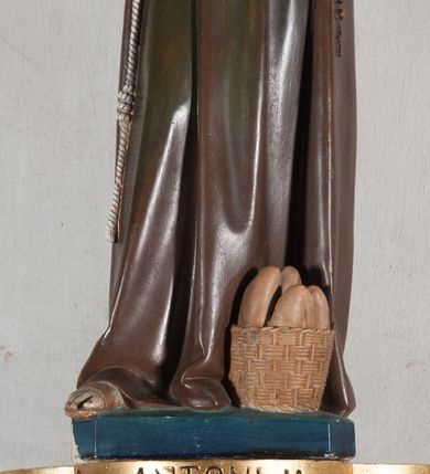 Zdjęcie nr 4: Rzeźba pełnoplastyczna, umieszczona na niewielkim prostopadłościennym postumencie o ściętych narożach oraz złoconej i srebrzonej konsoli utworzonej z liści akantu i róż. Święty ukazany całopostaciowo, frontalnie w pozycji stojącej. Antoni został przedstawiony jako młody mężczyzna o owalnej, pozbawionej zarostu twarzy i krótkich, brązowych włosach z tonsurą. Ma oczy wpatrzone przed siebie, wydatny, prosty nos, usta zamknięte. Postać w prawej ręce trzyma bochenek chleba, a na lewej podtrzymuje Dzieciątko Jezus. Święty ubrany jest w długi, ciemnobrązowy habit franciszkański. Do sznura z trzema węzłami ujmującego go w pasie przywiązany jest różaniec. Spod habitu widoczne stopy obute w sandały. U stóp ustawiony kosz z bochenkami chleba. Jasnowłose Dzieciątko Jezus ukazane w pozycji siedzącej z głową zwróconą w trzech czwartych w prawo. Prawą ręką wykonuje gest błogosławieństwa, lewą składa na piersi, podtrzymując kwiat białej lilii. Postać ubrana z białą tunikę ze złotą lamówką wokół dekoltu, rękawów oraz dolnej krawędzi szaty. Na konsoli, na której ustawiona jest rzeźba widnieje napis: ŚWIĘTY ANTONI MÓDL SIĘ ZA NAMI.