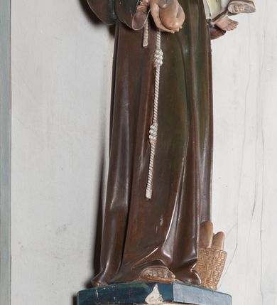 Zdjęcie nr 10: Rzeźba pełnoplastyczna, umieszczona na niewielkim prostopadłościennym postumencie o ściętych narożach oraz złoconej i srebrzonej konsoli utworzonej z liści akantu i róż. Święty ukazany całopostaciowo, frontalnie w pozycji stojącej. Antoni został przedstawiony jako młody mężczyzna o owalnej, pozbawionej zarostu twarzy i krótkich, brązowych włosach z tonsurą. Ma oczy wpatrzone przed siebie, wydatny, prosty nos, usta zamknięte. Postać w prawej ręce trzyma bochenek chleba, a na lewej podtrzymuje Dzieciątko Jezus. Święty ubrany jest w długi, ciemnobrązowy habit franciszkański. Do sznura z trzema węzłami ujmującego go w pasie przywiązany jest różaniec. Spod habitu widoczne stopy obute w sandały. U stóp ustawiony kosz z bochenkami chleba. Jasnowłose Dzieciątko Jezus ukazane w pozycji siedzącej z głową zwróconą w trzech czwartych w prawo. Prawą ręką wykonuje gest błogosławieństwa, lewą składa na piersi, podtrzymując kwiat białej lilii. Postać ubrana z białą tunikę ze złotą lamówką wokół dekoltu, rękawów oraz dolnej krawędzi szaty. Na konsoli, na której ustawiona jest rzeźba widnieje napis: ŚWIĘTY ANTONI MÓDL SIĘ ZA NAMI.