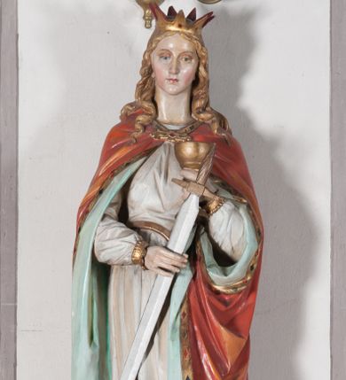 Zdjęcie nr 1: Rzeźba drążona, ustawiona na niewielkim, prostopadłościennym postumencie o ściętych narożach oraz rozbudowanej, neobarokowej konsoli. Święta ukazana frontalnie, w pozycji stojącej z kielichem w lewej dłoni i mieczem w prawej. Barbara została przedstawiona jako młoda, jasnowłosa kobieta o owalnej twarzy, dużych, niebieskich oczach i wydatnym nosie. Ubrana jest w strój królewski złożony z długiej, białej sukni ze złotymi elementami oraz czerwonego płaszcza z seledynowym podbiciem. Płaszcz jest spięty pod szyją ozdobną, złotą broszą. Na głowie postaci złota, wysadzana kamieniami szlachetnymi korona w typie otwartym. U jej stóp z lewej strony przedstawiona owalna wieża zwieńczona krenelażem. Konsola, na której ustawiona jest rzeźba wspierana jest przez postać jasnowłosego anioła w niebieskiej szacie trzymającego w rękach banderolę z napisem „Św(ięta) BARBARO Módl się za nami”. Anioł umieszczony na tle srebrzonego obłoku; banderola, skrzydła anioła oraz detale architektoniczne konsoli złocone.