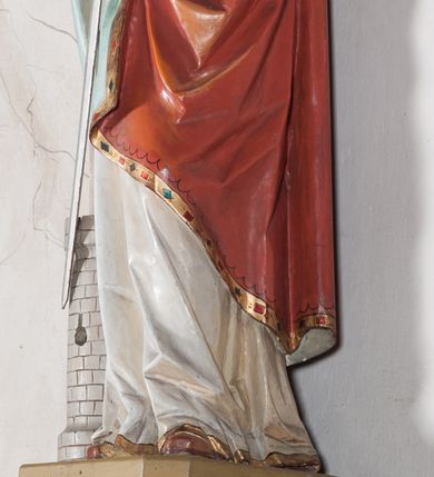 Zdjęcie nr 10: Rzeźba drążona, ustawiona na niewielkim, prostopadłościennym postumencie o ściętych narożach oraz rozbudowanej, neobarokowej konsoli. Święta ukazana frontalnie, w pozycji stojącej z kielichem w lewej dłoni i mieczem w prawej. Barbara została przedstawiona jako młoda, jasnowłosa kobieta o owalnej twarzy, dużych, niebieskich oczach i wydatnym nosie. Ubrana jest w strój królewski złożony z długiej, białej sukni ze złotymi elementami oraz czerwonego płaszcza z seledynowym podbiciem. Płaszcz jest spięty pod szyją ozdobną, złotą broszą. Na głowie postaci złota, wysadzana kamieniami szlachetnymi korona w typie otwartym. U jej stóp z lewej strony przedstawiona owalna wieża zwieńczona krenelażem. Konsola, na której ustawiona jest rzeźba wspierana jest przez postać jasnowłosego anioła w niebieskiej szacie trzymającego w rękach banderolę z napisem „Św(ięta) BARBARO Módl się za nami”. Anioł umieszczony na tle srebrzonego obłoku; banderola, skrzydła anioła oraz detale architektoniczne konsoli złocone.