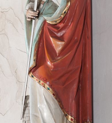 Zdjęcie nr 11: Rzeźba drążona, ustawiona na niewielkim, prostopadłościennym postumencie o ściętych narożach oraz rozbudowanej, neobarokowej konsoli. Święta ukazana frontalnie, w pozycji stojącej z kielichem w lewej dłoni i mieczem w prawej. Barbara została przedstawiona jako młoda, jasnowłosa kobieta o owalnej twarzy, dużych, niebieskich oczach i wydatnym nosie. Ubrana jest w strój królewski złożony z długiej, białej sukni ze złotymi elementami oraz czerwonego płaszcza z seledynowym podbiciem. Płaszcz jest spięty pod szyją ozdobną, złotą broszą. Na głowie postaci złota, wysadzana kamieniami szlachetnymi korona w typie otwartym. U jej stóp z lewej strony przedstawiona owalna wieża zwieńczona krenelażem. Konsola, na której ustawiona jest rzeźba wspierana jest przez postać jasnowłosego anioła w niebieskiej szacie trzymającego w rękach banderolę z napisem „Św(ięta) BARBARO Módl się za nami”. Anioł umieszczony na tle srebrzonego obłoku; banderola, skrzydła anioła oraz detale architektoniczne konsoli złocone.