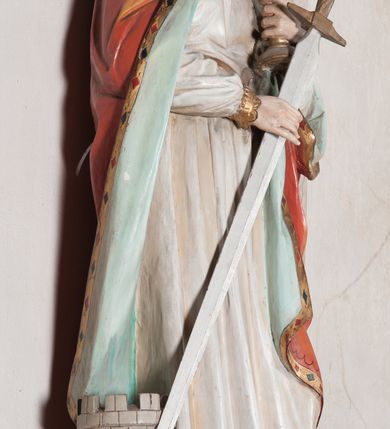 Zdjęcie nr 12: Rzeźba drążona, ustawiona na niewielkim, prostopadłościennym postumencie o ściętych narożach oraz rozbudowanej, neobarokowej konsoli. Święta ukazana frontalnie, w pozycji stojącej z kielichem w lewej dłoni i mieczem w prawej. Barbara została przedstawiona jako młoda, jasnowłosa kobieta o owalnej twarzy, dużych, niebieskich oczach i wydatnym nosie. Ubrana jest w strój królewski złożony z długiej, białej sukni ze złotymi elementami oraz czerwonego płaszcza z seledynowym podbiciem. Płaszcz jest spięty pod szyją ozdobną, złotą broszą. Na głowie postaci złota, wysadzana kamieniami szlachetnymi korona w typie otwartym. U jej stóp z lewej strony przedstawiona owalna wieża zwieńczona krenelażem. Konsola, na której ustawiona jest rzeźba wspierana jest przez postać jasnowłosego anioła w niebieskiej szacie trzymającego w rękach banderolę z napisem „Św(ięta) BARBARO Módl się za nami”. Anioł umieszczony na tle srebrzonego obłoku; banderola, skrzydła anioła oraz detale architektoniczne konsoli złocone.