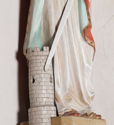 Zdjęcie nr 15: Rzeźba drążona, ustawiona na niewielkim, prostopadłościennym postumencie o ściętych narożach oraz rozbudowanej, neobarokowej konsoli. Święta ukazana frontalnie, w pozycji stojącej z kielichem w lewej dłoni i mieczem w prawej. Barbara została przedstawiona jako młoda, jasnowłosa kobieta o owalnej twarzy, dużych, niebieskich oczach i wydatnym nosie. Ubrana jest w strój królewski złożony z długiej, białej sukni ze złotymi elementami oraz czerwonego płaszcza z seledynowym podbiciem. Płaszcz jest spięty pod szyją ozdobną, złotą broszą. Na głowie postaci złota, wysadzana kamieniami szlachetnymi korona w typie otwartym. U jej stóp z lewej strony przedstawiona owalna wieża zwieńczona krenelażem. Konsola, na której ustawiona jest rzeźba wspierana jest przez postać jasnowłosego anioła w niebieskiej szacie trzymającego w rękach banderolę z napisem „Św(ięta) BARBARO Módl się za nami”. Anioł umieszczony na tle srebrzonego obłoku; banderola, skrzydła anioła oraz detale architektoniczne konsoli złocone.