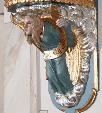 Zdjęcie nr 8: Rzeźba drążona, ustawiona na niewielkim, prostopadłościennym postumencie o ściętych narożach oraz rozbudowanej, neobarokowej konsoli. Święta ukazana frontalnie, w pozycji stojącej z kielichem w lewej dłoni i mieczem w prawej. Barbara została przedstawiona jako młoda, jasnowłosa kobieta o owalnej twarzy, dużych, niebieskich oczach i wydatnym nosie. Ubrana jest w strój królewski złożony z długiej, białej sukni ze złotymi elementami oraz czerwonego płaszcza z seledynowym podbiciem. Płaszcz jest spięty pod szyją ozdobną, złotą broszą. Na głowie postaci złota, wysadzana kamieniami szlachetnymi korona w typie otwartym. U jej stóp z lewej strony przedstawiona owalna wieża zwieńczona krenelażem. Konsola, na której ustawiona jest rzeźba wspierana jest przez postać jasnowłosego anioła w niebieskiej szacie trzymającego w rękach banderolę z napisem „Św(ięta) BARBARO Módl się za nami”. Anioł umieszczony na tle srebrzonego obłoku; banderola, skrzydła anioła oraz detale architektoniczne konsoli złocone.