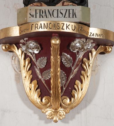 Zdjęcie nr 6: Rzeźba pełnoplastyczna, umieszczona na niewielkim prostopadłościennym postumencie o ściętych narożach oraz złoconej i srebrzonej konsoli utworzonej z liści akantu i róż. Święty ukazany całopostaciowo, frontalnie w pozycji stojącej. Franciszek został przedstawiony jako dojrzały mężczyzna o podłużnej, szczupłej twarzy i krótkich, brązowych włosach i brodzie. Ma duże, ciemne oczy wpatrzone przed siebie, wydatny ostro zakończony nos, usta zamknięte. Postać w prawej ręce trzyma krucyfiks, a lewą dłoń składa na piersi. Święty ubrany jest w długi, ciemnobrązowy habit franciszkański. Do sznura z trzema węzłami ujmującego go w pasie przywiązany jest różaniec. Spod habitu widoczne stopy obute w sandały. Na podstawie, na której ustawiona jest rzeźba widnieje napis „Św(ięty) FRANCISZEK”, na konsoli „ŚWIĘTY FRANCISZKU MÓDL SIĘ ZA NAMI”.