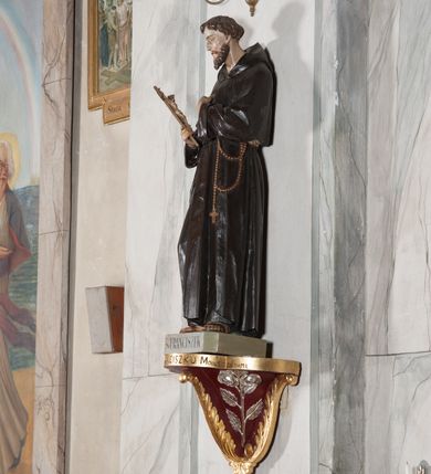 Zdjęcie nr 2: Rzeźba pełnoplastyczna, umieszczona na niewielkim prostopadłościennym postumencie o ściętych narożach oraz złoconej i srebrzonej konsoli utworzonej z liści akantu i róż. Święty ukazany całopostaciowo, frontalnie w pozycji stojącej. Franciszek został przedstawiony jako dojrzały mężczyzna o podłużnej, szczupłej twarzy i krótkich, brązowych włosach i brodzie. Ma duże, ciemne oczy wpatrzone przed siebie, wydatny ostro zakończony nos, usta zamknięte. Postać w prawej ręce trzyma krucyfiks, a lewą dłoń składa na piersi. Święty ubrany jest w długi, ciemnobrązowy habit franciszkański. Do sznura z trzema węzłami ujmującego go w pasie przywiązany jest różaniec. Spod habitu widoczne stopy obute w sandały. Na podstawie, na której ustawiona jest rzeźba widnieje napis „Św(ięty) FRANCISZEK”, na konsoli „ŚWIĘTY FRANCISZKU MÓDL SIĘ ZA NAMI”.