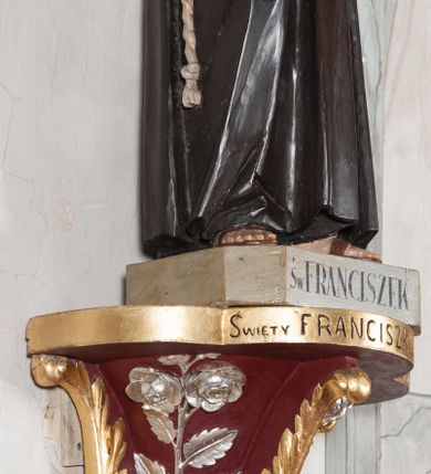 Zdjęcie nr 5: Rzeźba pełnoplastyczna, umieszczona na niewielkim prostopadłościennym postumencie o ściętych narożach oraz złoconej i srebrzonej konsoli utworzonej z liści akantu i róż. Święty ukazany całopostaciowo, frontalnie w pozycji stojącej. Franciszek został przedstawiony jako dojrzały mężczyzna o podłużnej, szczupłej twarzy i krótkich, brązowych włosach i brodzie. Ma duże, ciemne oczy wpatrzone przed siebie, wydatny ostro zakończony nos, usta zamknięte. Postać w prawej ręce trzyma krucyfiks, a lewą dłoń składa na piersi. Święty ubrany jest w długi, ciemnobrązowy habit franciszkański. Do sznura z trzema węzłami ujmującego go w pasie przywiązany jest różaniec. Spod habitu widoczne stopy obute w sandały. Na podstawie, na której ustawiona jest rzeźba widnieje napis „Św(ięty) FRANCISZEK”, na konsoli „ŚWIĘTY FRANCISZKU MÓDL SIĘ ZA NAMI”.