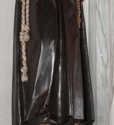 Zdjęcie nr 14: Rzeźba pełnoplastyczna, umieszczona na niewielkim prostopadłościennym postumencie o ściętych narożach oraz złoconej i srebrzonej konsoli utworzonej z liści akantu i róż. Święty ukazany całopostaciowo, frontalnie w pozycji stojącej. Franciszek został przedstawiony jako dojrzały mężczyzna o podłużnej, szczupłej twarzy i krótkich, brązowych włosach i brodzie. Ma duże, ciemne oczy wpatrzone przed siebie, wydatny ostro zakończony nos, usta zamknięte. Postać w prawej ręce trzyma krucyfiks, a lewą dłoń składa na piersi. Święty ubrany jest w długi, ciemnobrązowy habit franciszkański. Do sznura z trzema węzłami ujmującego go w pasie przywiązany jest różaniec. Spod habitu widoczne stopy obute w sandały. Na podstawie, na której ustawiona jest rzeźba widnieje napis „Św(ięty) FRANCISZEK”, na konsoli „ŚWIĘTY FRANCISZKU MÓDL SIĘ ZA NAMI”.