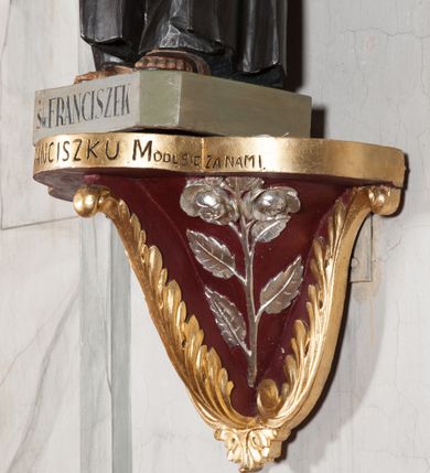 Zdjęcie nr 7: Rzeźba pełnoplastyczna, umieszczona na niewielkim prostopadłościennym postumencie o ściętych narożach oraz złoconej i srebrzonej konsoli utworzonej z liści akantu i róż. Święty ukazany całopostaciowo, frontalnie w pozycji stojącej. Franciszek został przedstawiony jako dojrzały mężczyzna o podłużnej, szczupłej twarzy i krótkich, brązowych włosach i brodzie. Ma duże, ciemne oczy wpatrzone przed siebie, wydatny ostro zakończony nos, usta zamknięte. Postać w prawej ręce trzyma krucyfiks, a lewą dłoń składa na piersi. Święty ubrany jest w długi, ciemnobrązowy habit franciszkański. Do sznura z trzema węzłami ujmującego go w pasie przywiązany jest różaniec. Spod habitu widoczne stopy obute w sandały. Na podstawie, na której ustawiona jest rzeźba widnieje napis „Św(ięty) FRANCISZEK”, na konsoli „ŚWIĘTY FRANCISZKU MÓDL SIĘ ZA NAMI”.