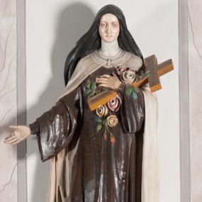 Zdjęcie nr 1: Rzeźba ustawiona na niewielkim, prostopadłościennym postumencie oraz rozbudowanej, neobarokowej konsoli. Święta ukazana frontalnie, w pozycji stojącej z prawą ręką wyciągniętą na bok oraz lewą złożoną na piersi, podtrzymującą krzyż opleciony różami. Teresa została ukazana jako młoda kobieta o owalnej twarzy, dużych oczach i wydatnym nosie. Ubrana jest w ciemnobrązowy, karmelitański habit z jasnym płaszczem. Konsola, na której ustawiona jest rzeźba wspierana jest przez postać jasnowłosego anioła w beżowej szacie trzymającego w rękach banderolę z napisem „Św(ięta) TERESO Módl się za nami&quot;. Anioł umieszczony na tle srebrzonego obłoku; banderola, skrzydła anioła oraz detale architektoniczne konsoli złocone.