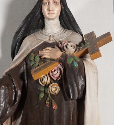 Zdjęcie nr 12: Rzeźba ustawiona na niewielkim, prostopadłościennym postumencie oraz rozbudowanej, neobarokowej konsoli. Święta ukazana frontalnie, w pozycji stojącej z prawą ręką wyciągniętą na bok oraz lewą złożoną na piersi, podtrzymującą krzyż opleciony różami. Teresa została ukazana jako młoda kobieta o owalnej twarzy, dużych oczach i wydatnym nosie. Ubrana jest w ciemnobrązowy, karmelitański habit z jasnym płaszczem. Konsola, na której ustawiona jest rzeźba wspierana jest przez postać jasnowłosego anioła w beżowej szacie trzymającego w rękach banderolę z napisem „Św(ięta) TERESO Módl się za nami". Anioł umieszczony na tle srebrzonego obłoku; banderola, skrzydła anioła oraz detale architektoniczne konsoli złocone.