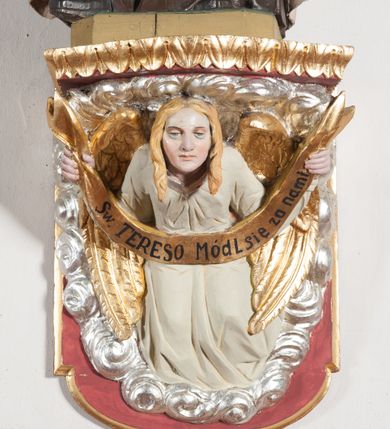 Zdjęcie nr 13: Rzeźba ustawiona na niewielkim, prostopadłościennym postumencie oraz rozbudowanej, neobarokowej konsoli. Święta ukazana frontalnie, w pozycji stojącej z prawą ręką wyciągniętą na bok oraz lewą złożoną na piersi, podtrzymującą krzyż opleciony różami. Teresa została ukazana jako młoda kobieta o owalnej twarzy, dużych oczach i wydatnym nosie. Ubrana jest w ciemnobrązowy, karmelitański habit z jasnym płaszczem. Konsola, na której ustawiona jest rzeźba wspierana jest przez postać jasnowłosego anioła w beżowej szacie trzymającego w rękach banderolę z napisem „Św(ięta) TERESO Módl się za nami&quot;. Anioł umieszczony na tle srebrzonego obłoku; banderola, skrzydła anioła oraz detale architektoniczne konsoli złocone.