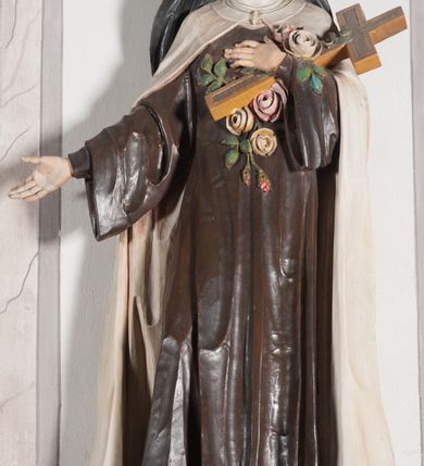 Zdjęcie nr 8: Rzeźba ustawiona na niewielkim, prostopadłościennym postumencie oraz rozbudowanej, neobarokowej konsoli. Święta ukazana frontalnie, w pozycji stojącej z prawą ręką wyciągniętą na bok oraz lewą złożoną na piersi, podtrzymującą krzyż opleciony różami. Teresa została ukazana jako młoda kobieta o owalnej twarzy, dużych oczach i wydatnym nosie. Ubrana jest w ciemnobrązowy, karmelitański habit z jasnym płaszczem. Konsola, na której ustawiona jest rzeźba wspierana jest przez postać jasnowłosego anioła w beżowej szacie trzymającego w rękach banderolę z napisem „Św(ięta) TERESO Módl się za nami". Anioł umieszczony na tle srebrzonego obłoku; banderola, skrzydła anioła oraz detale architektoniczne konsoli złocone.