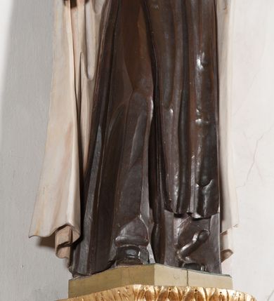 Zdjęcie nr 6: Rzeźba ustawiona na niewielkim, prostopadłościennym postumencie oraz rozbudowanej, neobarokowej konsoli. Święta ukazana frontalnie, w pozycji stojącej z prawą ręką wyciągniętą na bok oraz lewą złożoną na piersi, podtrzymującą krzyż opleciony różami. Teresa została ukazana jako młoda kobieta o owalnej twarzy, dużych oczach i wydatnym nosie. Ubrana jest w ciemnobrązowy, karmelitański habit z jasnym płaszczem. Konsola, na której ustawiona jest rzeźba wspierana jest przez postać jasnowłosego anioła w beżowej szacie trzymającego w rękach banderolę z napisem „Św(ięta) TERESO Módl się za nami". Anioł umieszczony na tle srebrzonego obłoku; banderola, skrzydła anioła oraz detale architektoniczne konsoli złocone.