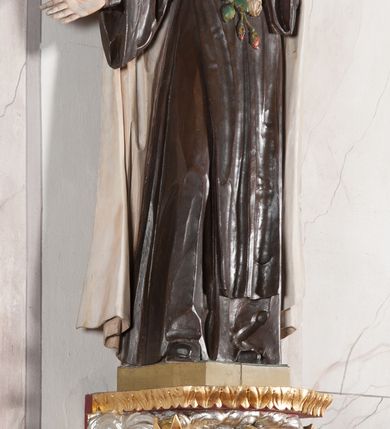 Zdjęcie nr 2: Rzeźba ustawiona na niewielkim, prostopadłościennym postumencie oraz rozbudowanej, neobarokowej konsoli. Święta ukazana frontalnie, w pozycji stojącej z prawą ręką wyciągniętą na bok oraz lewą złożoną na piersi, podtrzymującą krzyż opleciony różami. Teresa została ukazana jako młoda kobieta o owalnej twarzy, dużych oczach i wydatnym nosie. Ubrana jest w ciemnobrązowy, karmelitański habit z jasnym płaszczem. Konsola, na której ustawiona jest rzeźba wspierana jest przez postać jasnowłosego anioła w beżowej szacie trzymającego w rękach banderolę z napisem „Św(ięta) TERESO Módl się za nami". Anioł umieszczony na tle srebrzonego obłoku; banderola, skrzydła anioła oraz detale architektoniczne konsoli złocone.