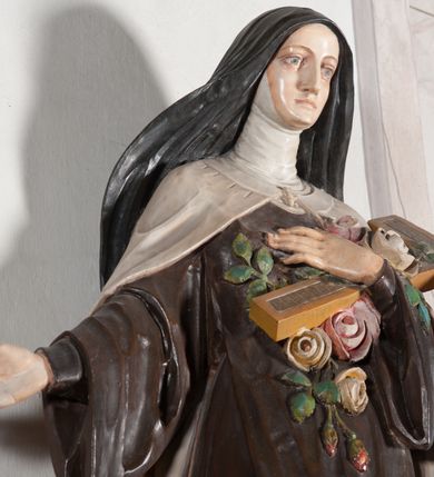 Zdjęcie nr 10: Rzeźba ustawiona na niewielkim, prostopadłościennym postumencie oraz rozbudowanej, neobarokowej konsoli. Święta ukazana frontalnie, w pozycji stojącej z prawą ręką wyciągniętą na bok oraz lewą złożoną na piersi, podtrzymującą krzyż opleciony różami. Teresa została ukazana jako młoda kobieta o owalnej twarzy, dużych oczach i wydatnym nosie. Ubrana jest w ciemnobrązowy, karmelitański habit z jasnym płaszczem. Konsola, na której ustawiona jest rzeźba wspierana jest przez postać jasnowłosego anioła w beżowej szacie trzymającego w rękach banderolę z napisem „Św(ięta) TERESO Módl się za nami". Anioł umieszczony na tle srebrzonego obłoku; banderola, skrzydła anioła oraz detale architektoniczne konsoli złocone.