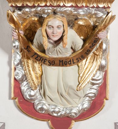 Zdjęcie nr 3: Rzeźba ustawiona na niewielkim, prostopadłościennym postumencie oraz rozbudowanej, neobarokowej konsoli. Święta ukazana frontalnie, w pozycji stojącej z prawą ręką wyciągniętą na bok oraz lewą złożoną na piersi, podtrzymującą krzyż opleciony różami. Teresa została ukazana jako młoda kobieta o owalnej twarzy, dużych oczach i wydatnym nosie. Ubrana jest w ciemnobrązowy, karmelitański habit z jasnym płaszczem. Konsola, na której ustawiona jest rzeźba wspierana jest przez postać jasnowłosego anioła w beżowej szacie trzymającego w rękach banderolę z napisem „Św(ięta) TERESO Módl się za nami". Anioł umieszczony na tle srebrzonego obłoku; banderola, skrzydła anioła oraz detale architektoniczne konsoli złocone.