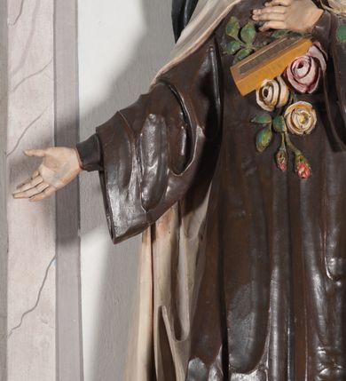 Zdjęcie nr 11: Rzeźba ustawiona na niewielkim, prostopadłościennym postumencie oraz rozbudowanej, neobarokowej konsoli. Święta ukazana frontalnie, w pozycji stojącej z prawą ręką wyciągniętą na bok oraz lewą złożoną na piersi, podtrzymującą krzyż opleciony różami. Teresa została ukazana jako młoda kobieta o owalnej twarzy, dużych oczach i wydatnym nosie. Ubrana jest w ciemnobrązowy, karmelitański habit z jasnym płaszczem. Konsola, na której ustawiona jest rzeźba wspierana jest przez postać jasnowłosego anioła w beżowej szacie trzymającego w rękach banderolę z napisem „Św(ięta) TERESO Módl się za nami". Anioł umieszczony na tle srebrzonego obłoku; banderola, skrzydła anioła oraz detale architektoniczne konsoli złocone.