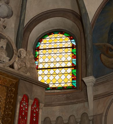 Zdjęcie nr 2: Okno w kształcie stojącego prostokąta zamkniętego półkoliście.  Prosta kompozycja geometryczna w postaci ukośnej kratki utworzonej z naprzemiennie ułożonych białych i żółtych rombów. Całość ujęta geometryczną bordiurą w kolorach zielony, pomarańczowym oraz odcieniach czerwieni.