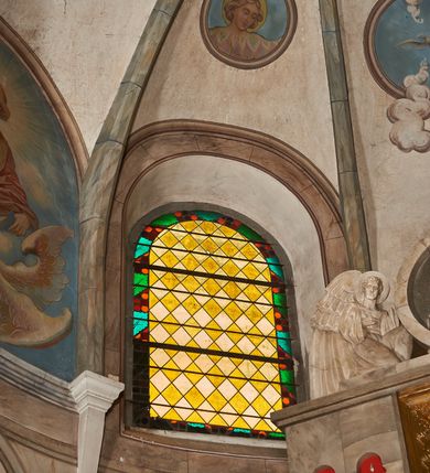 Zdjęcie nr 1: Okno w kształcie stojącego prostokąta zamkniętego półkoliście.  Prosta kompozycja geometryczna w postaci ukośnej kratki utworzonej z naprzemiennie ułożonych białych i żółtych rombów. Całość ujęta geometryczną bordiurą w kolorach zielony, pomarańczowym oraz odcieniach czerwieni.