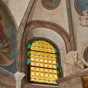 Zdjęcie nr 1: Okno w kształcie stojącego prostokąta zamkniętego półkoliście.  Prosta kompozycja geometryczna w postaci ukośnej kratki utworzonej z naprzemiennie ułożonych białych i żółtych rombów. Całość ujęta geometryczną bordiurą w kolorach zielony, pomarańczowym oraz odcieniach czerwieni.