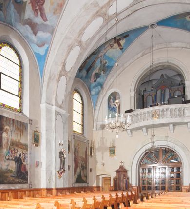 Zdjęcie nr 18: Prezbiterium: 
Sklepienie krzyżowo-żebrowe, dwuprzęsłowe. Pola sklepienne malowane są na kolor beżowy. Od strony wschodniej z centralnie umieszczonymi trzema kolistymi medalionami, mieszczącymi przedstawienie gołębicy Ducha Świętego w obłokach na błękitnym tle (w medalionie centralnym) oraz przeciwstawnie ustawione popiersia dwóch serafinów przedstawionych jako jasnowłosi młodzieńcy w pastelowych szatach, z nimbem wokół głowy (w medalionach skrajnych). W ścianach tarczowych umieszczone wizerunki czterech ewangelistów wraz z ich atrybutami. W pierwszym od wschodu przęśle przedstawienie św. Jana z orłem (po lewej od ołtarza) i św. Mateusza z aniołem (po prawej). W przęśle drugim wizerunek św. Łukasza z uskrzydlonym bykiem (po lewej) i św. Marka z uskrzydlonym lwem (po prawej). Ewangeliści ukazani w popiersiu, na tle błękitnego nieba, obłoków i promienistego nimbu. W dolnej części każdej z kompozycji przedstawione symbole każdego z ewangelistów. Święty Jan ukazany jako młody, jasnowłosy mężczyzna z głową zwróconą w prawo. Ubrany jest w jasnozieloną szatę oraz luźno przerzucony przez ramię różowy płaszcz. W prawym ręku trzyma pióro, w lewym księgę. Świętego Mateusza przedstawiono jako starszego, siwowłosego i brodatego mężczyznę z głową zwróconą w lewo. Ubrany jest w złotą szatę oraz błękitny płaszcz. Swoją lewą rękę składa na piersi, prawą wskazuje w dół. Święty Łukasz przedstawiony jest jako starszy siwowłosy i brodaty mężczyzna ukazany w dynamicznej pozie – z głową z profilu zwróconą w lewo oraz lewą ręką uniesioną do góry. Postać ubrana jest w jasnofioletową szatę oraz błękitny płaszcz. Świętego Marka przedstawiono z ciałem zwróconym w trzech czwartych w prawo i głową ujętą z profilu. Ewangelistę ukazano jako dojrzałego, brodatego mężczyznę o ostrych rysach. Ubrany jest w różową szatę oraz błękitny płaszcz z jasnozieloną podszewką. W swoim prawym ręku trzyma pióro. Elementy architektoniczne takie jak żebra sklepienne, obramienia okien i arkadowych nisz oraz gzyms przebiegający na linii parapetów okiennych są polichromowane w odcieniach szarości z efektem marmoryzacji naśladującej kolorystykę alabastrowych elementów wyposażenia kościoła.
Arkada i ściana tęczowa: Na ścianie tęczowej znajduje się wielopostaciowa, figuralna kompozycja z umieszczonym w centrum przedstawieniem Matki Boskiej Częstochowskiej unoszącej się na skrzydłach orła białego. Przedstawienie to jest flankowane przez dwie grupy świętych. Popiersie Marii z Dzieciątkiem ukazano na tle obłoków oraz promienistej glorii oraz ujęto wieńcem dwunastu gwiazd. Z lewej strony kompozycji ukazano sześcioosobową grupę klęczących świętych, zwracających się w kierunku Marii. Od lewej przedstawiony św. Andrzej Bobola z rękoma skrzyżowanymi na piersi, św. Wojciech w infule i paliuszu szeroko rozpościerający ramiona, św. Kinga w brązowym habicie klaryski z koroną na głowie, błogosławiona Bronisława w jasnoróżowym habicie, z krucyfiksem w dłoni, św. Stanisław biskup i męczennik z pastorałem w dłoni oraz infule na głowie i św. Jadwiga Królowa przedstawiona w złotej koronie i gronostajowym płaszczu. Sześcioosobową grupę świętych ukazanych z prawej strony kompozycji od lewej otwiera postać św. Jana Kantego przedstawionego w birecie oraz obszernym, czerwonym płaszczu z gronostajowym kołnierzem. Za nim przedstawiony św. Stanisław Kostka z rękoma skrzyżowanymi na piersi, następnie św. Jacek w habicie dominikańskim z monstrancją w ręku, błogosławiony Wincenty Kadłubek w stroju biskupim, św. Kazimierz Królewicz w mitrze książęcej oraz czerwonym płaszczu podbitym gronostajem i błogosławiona Salomea przedstawiona w brązowym habicie klaryski, z mitrą na głowie.  
Nawa główna: W nawie sklepienie kolebkowe z lunetami, trójprzęsłowe. Pola sklepienne malowane na kolor jasnobeżowy. W każdym z przęseł, w lunetach sceny figuralne, tworzące cykl ikonograficzny obrazujący sceny z życia Chrystusa i Marii. Wszystkie sceny umieszczone na tle nieba i obłoków. Cykl otwiera scena Zwiastowania umieszczona w pierwszym od prezbiterium przęśle sklepienia po lewej (północnej) stronie. W obłokach ukazani Maria (z prawej) oraz archanioł Gabriel (z lewej), ponad którymi unosi się gołębica Ducha Świętego otoczona świetlistą glorią. Maria przedstawiona jako młoda, jasnowłosa kobieta z ciałem zwróconym w prawo. Głowa otoczona nimbem, pochylona do dołu, prawa ręka złożona na piersi. Postać ubrana jest w białą suknię oraz błękitny płaszcz. Archanioł Gabriel ukazany z profilu w dynamicznej pozie z prawą ręką uniesioną do góry i lewą wyciągniętą do przodu, w której trzyma kwiat lilii. Anioł ubrany jest w długą, jasnoróżową szatę. W kolejnym przęśle przedstawiono Boże Narodzenie. W dolnej części kompozycji ukazani św. Józef (z lewej) i Maria (z prawej) klęczący w obłokach, obok żłóbka z Dzieciątkiem Jezus. Józef ukazany jako dojrzały, brodaty mężczyzna w żółtej szacie i zielonym płaszczu, trzymający w prawej ręce kwiat lilii. Maria przedstawiona jako młoda kobieta z dłońmi złożonymi w geście modlitwy, ubrana w jasnoróżową szatę oraz błękitny płaszcz, którego poła nakrywa jej głowę. Dzieciątko Jezus przepasane białą materią, ukazane frontalnie z rękami wyciągniętymi do góry. W górnej części kompozycji przedstawiony frontalnie długowłosy anioł w jasnozielonej szacie trzymający oburącz banderolę z napisem „GLORIA IN EXCELSIS DEO”. W trzecim przęśle scena Modlitwy w Ogrójcu. Z lewej strony kompozycji przedstawiony z profilu, klęczący Chrystus unoszący do góry ręce złożone w geście modlitwy. Postać ubrana w długą, białą szatę oraz luźno przerzucony przez ramię jasnobłękitny płaszcz. Z prawej strony kompozycji, naprzeciw Chrystusa ukazujący się w obłokach anioł trzymający złoty kielich. Anioł przedstawiony jako długowłosa, młodzieńcza postać ubrana w biało-zielone szaty. Wokół obu postaci rama utworzona z ciemnoszarych, skłębionych obłoków. Kolejne przedstawienia figuralne znajdują się w prawej (południowej) części sklepienia. W pierwszym od strony chóru przęśle przedstawiono Ukrzyżowanie. W centrum kompozycji ukrzyżowany Chrystus z ramionami uniesionymi do góry, tworzącymi linię litery „V”, uniesioną głową w koronie cierniowej oraz stopami przebitymi jednym gwoździem. Postać przepasana długim, białym perizonium. U stóp krzyża ukazani w dynamicznych pozach, z rękoma wzniesionymi do góry św. Jan Ewangelista (z lewej) oraz Maria (z prawej). Święty Jan ubrany w zieloną szatę oraz rozwiany żółto-różowy płaszcz. Maria ukazana w jasnoróżowej sukni oraz granatowo-zielonym płaszczu i chuście na głowie. W kolejnym przęśle przedstawiono scenę Zmartwychwstania. W centrum kompozycji ukazany frontalnie, całopostaciowo zmartwychwstały Chrystus w obszernym, układającym się w dynamicznie załamujące się fałdy, czerwonym płaszczu. Jezus prawą, uniesioną do góry ręką błogosławi, w lewej trzyma chorągiew paschalną. W dolnej części kompozycji przedstawiony anioł w błękitnej szacie podtrzymujący wieko grobowca. W ostatnim przęśle znajduje się scena Wniebowzięcia Marii. W centrum kompozycji, na jasnożółtym tle frontalnie przedstawiona, unosząca się ponad obłokami Maria. Postać ukazana jako młoda kobieta z głową otoczoną wieńcem z gwiazd i rękami uniesionymi do góry. Ubrana jest w jasnoróżową suknię oraz spięty pod szyją, błękitny płaszcz. W dolnej części kompozycji dwa anioły ukazane w dynamicznych pozach. Anioł z lewej strony w zielonej sukni, anioł z prawej w jasnoróżowej. Na ścianie zachodniej kościoła, na wysokości parapetu chóru muzycznego dwie prostokątne płyciny zamknięte łukiem półokrągłym. Płycina z lewej mieści przedstawienie św. Cecylii grającej na organach, płycina z prawej wizerunek króla Dawida grającego na harfie. Pasy gurtów sklepiennych w nawie dekorowane są geometryczną dekoracją z malowanymi pseudopłycinami w kształcie poziomego prostokąta o krótszych bokach zamkniętych łukiem półkolistym z uskokiem. Filary artykułujące nawę, parapet chóru muzycznego oraz gzyms na ścianie zachodniej świątyni polichromowane w odcieniach szarości z efektem marmoryzacji naśladującej kolorystykę alabastrowych elementów wyposażenia kościoła. 
