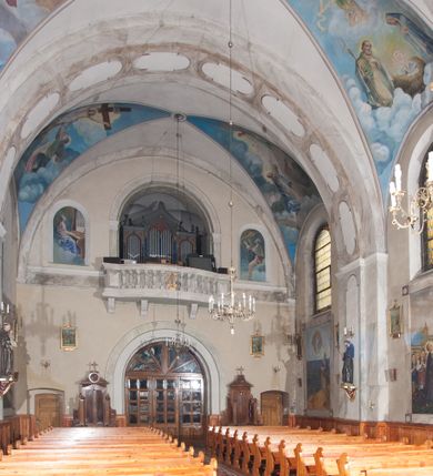 Zdjęcie nr 17: Prezbiterium: 
Sklepienie krzyżowo-żebrowe, dwuprzęsłowe. Pola sklepienne malowane są na kolor beżowy. Od strony wschodniej z centralnie umieszczonymi trzema kolistymi medalionami, mieszczącymi przedstawienie gołębicy Ducha Świętego w obłokach na błękitnym tle (w medalionie centralnym) oraz przeciwstawnie ustawione popiersia dwóch serafinów przedstawionych jako jasnowłosi młodzieńcy w pastelowych szatach, z nimbem wokół głowy (w medalionach skrajnych). W ścianach tarczowych umieszczone wizerunki czterech ewangelistów wraz z ich atrybutami. W pierwszym od wschodu przęśle przedstawienie św. Jana z orłem (po lewej od ołtarza) i św. Mateusza z aniołem (po prawej). W przęśle drugim wizerunek św. Łukasza z uskrzydlonym bykiem (po lewej) i św. Marka z uskrzydlonym lwem (po prawej). Ewangeliści ukazani w popiersiu, na tle błękitnego nieba, obłoków i promienistego nimbu. W dolnej części każdej z kompozycji przedstawione symbole każdego z ewangelistów. Święty Jan ukazany jako młody, jasnowłosy mężczyzna z głową zwróconą w prawo. Ubrany jest w jasnozieloną szatę oraz luźno przerzucony przez ramię różowy płaszcz. W prawym ręku trzyma pióro, w lewym księgę. Świętego Mateusza przedstawiono jako starszego, siwowłosego i brodatego mężczyznę z głową zwróconą w lewo. Ubrany jest w złotą szatę oraz błękitny płaszcz. Swoją lewą rękę składa na piersi, prawą wskazuje w dół. Święty Łukasz przedstawiony jest jako starszy siwowłosy i brodaty mężczyzna ukazany w dynamicznej pozie – z głową z profilu zwróconą w lewo oraz lewą ręką uniesioną do góry. Postać ubrana jest w jasnofioletową szatę oraz błękitny płaszcz. Świętego Marka przedstawiono z ciałem zwróconym w trzech czwartych w prawo i głową ujętą z profilu. Ewangelistę ukazano jako dojrzałego, brodatego mężczyznę o ostrych rysach. Ubrany jest w różową szatę oraz błękitny płaszcz z jasnozieloną podszewką. W swoim prawym ręku trzyma pióro. Elementy architektoniczne takie jak żebra sklepienne, obramienia okien i arkadowych nisz oraz gzyms przebiegający na linii parapetów okiennych są polichromowane w odcieniach szarości z efektem marmoryzacji naśladującej kolorystykę alabastrowych elementów wyposażenia kościoła.
Arkada i ściana tęczowa: Na ścianie tęczowej znajduje się wielopostaciowa, figuralna kompozycja z umieszczonym w centrum przedstawieniem Matki Boskiej Częstochowskiej unoszącej się na skrzydłach orła białego. Przedstawienie to jest flankowane przez dwie grupy świętych. Popiersie Marii z Dzieciątkiem ukazano na tle obłoków oraz promienistej glorii oraz ujęto wieńcem dwunastu gwiazd. Z lewej strony kompozycji ukazano sześcioosobową grupę klęczących świętych, zwracających się w kierunku Marii. Od lewej przedstawiony św. Andrzej Bobola z rękoma skrzyżowanymi na piersi, św. Wojciech w infule i paliuszu szeroko rozpościerający ramiona, św. Kinga w brązowym habicie klaryski z koroną na głowie, błogosławiona Bronisława w jasnoróżowym habicie, z krucyfiksem w dłoni, św. Stanisław biskup i męczennik z pastorałem w dłoni oraz infule na głowie i św. Jadwiga Królowa przedstawiona w złotej koronie i gronostajowym płaszczu. Sześcioosobową grupę świętych ukazanych z prawej strony kompozycji od lewej otwiera postać św. Jana Kantego przedstawionego w birecie oraz obszernym, czerwonym płaszczu z gronostajowym kołnierzem. Za nim przedstawiony św. Stanisław Kostka z rękoma skrzyżowanymi na piersi, następnie św. Jacek w habicie dominikańskim z monstrancją w ręku, błogosławiony Wincenty Kadłubek w stroju biskupim, św. Kazimierz Królewicz w mitrze książęcej oraz czerwonym płaszczu podbitym gronostajem i błogosławiona Salomea przedstawiona w brązowym habicie klaryski, z mitrą na głowie.  
Nawa główna: W nawie sklepienie kolebkowe z lunetami, trójprzęsłowe. Pola sklepienne malowane na kolor jasnobeżowy. W każdym z przęseł, w lunetach sceny figuralne, tworzące cykl ikonograficzny obrazujący sceny z życia Chrystusa i Marii. Wszystkie sceny umieszczone na tle nieba i obłoków. Cykl otwiera scena Zwiastowania umieszczona w pierwszym od prezbiterium przęśle sklepienia po lewej (północnej) stronie. W obłokach ukazani Maria (z prawej) oraz archanioł Gabriel (z lewej), ponad którymi unosi się gołębica Ducha Świętego otoczona świetlistą glorią. Maria przedstawiona jako młoda, jasnowłosa kobieta z ciałem zwróconym w prawo. Głowa otoczona nimbem, pochylona do dołu, prawa ręka złożona na piersi. Postać ubrana jest w białą suknię oraz błękitny płaszcz. Archanioł Gabriel ukazany z profilu w dynamicznej pozie z prawą ręką uniesioną do góry i lewą wyciągniętą do przodu, w której trzyma kwiat lilii. Anioł ubrany jest w długą, jasnoróżową szatę. W kolejnym przęśle przedstawiono Boże Narodzenie. W dolnej części kompozycji ukazani św. Józef (z lewej) i Maria (z prawej) klęczący w obłokach, obok żłóbka z Dzieciątkiem Jezus. Józef ukazany jako dojrzały, brodaty mężczyzna w żółtej szacie i zielonym płaszczu, trzymający w prawej ręce kwiat lilii. Maria przedstawiona jako młoda kobieta z dłońmi złożonymi w geście modlitwy, ubrana w jasnoróżową szatę oraz błękitny płaszcz, którego poła nakrywa jej głowę. Dzieciątko Jezus przepasane białą materią, ukazane frontalnie z rękami wyciągniętymi do góry. W górnej części kompozycji przedstawiony frontalnie długowłosy anioł w jasnozielonej szacie trzymający oburącz banderolę z napisem „GLORIA IN EXCELSIS DEO”. W trzecim przęśle scena Modlitwy w Ogrójcu. Z lewej strony kompozycji przedstawiony z profilu, klęczący Chrystus unoszący do góry ręce złożone w geście modlitwy. Postać ubrana w długą, białą szatę oraz luźno przerzucony przez ramię jasnobłękitny płaszcz. Z prawej strony kompozycji, naprzeciw Chrystusa ukazujący się w obłokach anioł trzymający złoty kielich. Anioł przedstawiony jako długowłosa, młodzieńcza postać ubrana w biało-zielone szaty. Wokół obu postaci rama utworzona z ciemnoszarych, skłębionych obłoków. Kolejne przedstawienia figuralne znajdują się w prawej (południowej) części sklepienia. W pierwszym od strony chóru przęśle przedstawiono Ukrzyżowanie. W centrum kompozycji ukrzyżowany Chrystus z ramionami uniesionymi do góry, tworzącymi linię litery „V”, uniesioną głową w koronie cierniowej oraz stopami przebitymi jednym gwoździem. Postać przepasana długim, białym perizonium. U stóp krzyża ukazani w dynamicznych pozach, z rękoma wzniesionymi do góry św. Jan Ewangelista (z lewej) oraz Maria (z prawej). Święty Jan ubrany w zieloną szatę oraz rozwiany żółto-różowy płaszcz. Maria ukazana w jasnoróżowej sukni oraz granatowo-zielonym płaszczu i chuście na głowie. W kolejnym przęśle przedstawiono scenę Zmartwychwstania. W centrum kompozycji ukazany frontalnie, całopostaciowo zmartwychwstały Chrystus w obszernym, układającym się w dynamicznie załamujące się fałdy, czerwonym płaszczu. Jezus prawą, uniesioną do góry ręką błogosławi, w lewej trzyma chorągiew paschalną. W dolnej części kompozycji przedstawiony anioł w błękitnej szacie podtrzymujący wieko grobowca. W ostatnim przęśle znajduje się scena Wniebowzięcia Marii. W centrum kompozycji, na jasnożółtym tle frontalnie przedstawiona, unosząca się ponad obłokami Maria. Postać ukazana jako młoda kobieta z głową otoczoną wieńcem z gwiazd i rękami uniesionymi do góry. Ubrana jest w jasnoróżową suknię oraz spięty pod szyją, błękitny płaszcz. W dolnej części kompozycji dwa anioły ukazane w dynamicznych pozach. Anioł z lewej strony w zielonej sukni, anioł z prawej w jasnoróżowej. Na ścianie zachodniej kościoła, na wysokości parapetu chóru muzycznego dwie prostokątne płyciny zamknięte łukiem półokrągłym. Płycina z lewej mieści przedstawienie św. Cecylii grającej na organach, płycina z prawej wizerunek króla Dawida grającego na harfie. Pasy gurtów sklepiennych w nawie dekorowane są geometryczną dekoracją z malowanymi pseudopłycinami w kształcie poziomego prostokąta o krótszych bokach zamkniętych łukiem półkolistym z uskokiem. Filary artykułujące nawę, parapet chóru muzycznego oraz gzyms na ścianie zachodniej świątyni polichromowane w odcieniach szarości z efektem marmoryzacji naśladującej kolorystykę alabastrowych elementów wyposażenia kościoła. 
