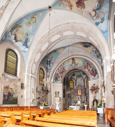 Zdjęcie nr 16: Prezbiterium: 
Sklepienie krzyżowo-żebrowe, dwuprzęsłowe. Pola sklepienne malowane są na kolor beżowy. Od strony wschodniej z centralnie umieszczonymi trzema kolistymi medalionami, mieszczącymi przedstawienie gołębicy Ducha Świętego w obłokach na błękitnym tle (w medalionie centralnym) oraz przeciwstawnie ustawione popiersia dwóch serafinów przedstawionych jako jasnowłosi młodzieńcy w pastelowych szatach, z nimbem wokół głowy (w medalionach skrajnych). W ścianach tarczowych umieszczone wizerunki czterech ewangelistów wraz z ich atrybutami. W pierwszym od wschodu przęśle przedstawienie św. Jana z orłem (po lewej od ołtarza) i św. Mateusza z aniołem (po prawej). W przęśle drugim wizerunek św. Łukasza z uskrzydlonym bykiem (po lewej) i św. Marka z uskrzydlonym lwem (po prawej). Ewangeliści ukazani w popiersiu, na tle błękitnego nieba, obłoków i promienistego nimbu. W dolnej części każdej z kompozycji przedstawione symbole każdego z ewangelistów. Święty Jan ukazany jako młody, jasnowłosy mężczyzna z głową zwróconą w prawo. Ubrany jest w jasnozieloną szatę oraz luźno przerzucony przez ramię różowy płaszcz. W prawym ręku trzyma pióro, w lewym księgę. Świętego Mateusza przedstawiono jako starszego, siwowłosego i brodatego mężczyznę z głową zwróconą w lewo. Ubrany jest w złotą szatę oraz błękitny płaszcz. Swoją lewą rękę składa na piersi, prawą wskazuje w dół. Święty Łukasz przedstawiony jest jako starszy siwowłosy i brodaty mężczyzna ukazany w dynamicznej pozie – z głową z profilu zwróconą w lewo oraz lewą ręką uniesioną do góry. Postać ubrana jest w jasnofioletową szatę oraz błękitny płaszcz. Świętego Marka przedstawiono z ciałem zwróconym w trzech czwartych w prawo i głową ujętą z profilu. Ewangelistę ukazano jako dojrzałego, brodatego mężczyznę o ostrych rysach. Ubrany jest w różową szatę oraz błękitny płaszcz z jasnozieloną podszewką. W swoim prawym ręku trzyma pióro. Elementy architektoniczne takie jak żebra sklepienne, obramienia okien i arkadowych nisz oraz gzyms przebiegający na linii parapetów okiennych są polichromowane w odcieniach szarości z efektem marmoryzacji naśladującej kolorystykę alabastrowych elementów wyposażenia kościoła.
Arkada i ściana tęczowa: Na ścianie tęczowej znajduje się wielopostaciowa, figuralna kompozycja z umieszczonym w centrum przedstawieniem Matki Boskiej Częstochowskiej unoszącej się na skrzydłach orła białego. Przedstawienie to jest flankowane przez dwie grupy świętych. Popiersie Marii z Dzieciątkiem ukazano na tle obłoków oraz promienistej glorii oraz ujęto wieńcem dwunastu gwiazd. Z lewej strony kompozycji ukazano sześcioosobową grupę klęczących świętych, zwracających się w kierunku Marii. Od lewej przedstawiony św. Andrzej Bobola z rękoma skrzyżowanymi na piersi, św. Wojciech w infule i paliuszu szeroko rozpościerający ramiona, św. Kinga w brązowym habicie klaryski z koroną na głowie, błogosławiona Bronisława w jasnoróżowym habicie, z krucyfiksem w dłoni, św. Stanisław biskup i męczennik z pastorałem w dłoni oraz infule na głowie i św. Jadwiga Królowa przedstawiona w złotej koronie i gronostajowym płaszczu. Sześcioosobową grupę świętych ukazanych z prawej strony kompozycji od lewej otwiera postać św. Jana Kantego przedstawionego w birecie oraz obszernym, czerwonym płaszczu z gronostajowym kołnierzem. Za nim przedstawiony św. Stanisław Kostka z rękoma skrzyżowanymi na piersi, następnie św. Jacek w habicie dominikańskim z monstrancją w ręku, błogosławiony Wincenty Kadłubek w stroju biskupim, św. Kazimierz Królewicz w mitrze książęcej oraz czerwonym płaszczu podbitym gronostajem i błogosławiona Salomea przedstawiona w brązowym habicie klaryski, z mitrą na głowie.  
Nawa główna: W nawie sklepienie kolebkowe z lunetami, trójprzęsłowe. Pola sklepienne malowane na kolor jasnobeżowy. W każdym z przęseł, w lunetach sceny figuralne, tworzące cykl ikonograficzny obrazujący sceny z życia Chrystusa i Marii. Wszystkie sceny umieszczone na tle nieba i obłoków. Cykl otwiera scena Zwiastowania umieszczona w pierwszym od prezbiterium przęśle sklepienia po lewej (północnej) stronie. W obłokach ukazani Maria (z prawej) oraz archanioł Gabriel (z lewej), ponad którymi unosi się gołębica Ducha Świętego otoczona świetlistą glorią. Maria przedstawiona jako młoda, jasnowłosa kobieta z ciałem zwróconym w prawo. Głowa otoczona nimbem, pochylona do dołu, prawa ręka złożona na piersi. Postać ubrana jest w białą suknię oraz błękitny płaszcz. Archanioł Gabriel ukazany z profilu w dynamicznej pozie z prawą ręką uniesioną do góry i lewą wyciągniętą do przodu, w której trzyma kwiat lilii. Anioł ubrany jest w długą, jasnoróżową szatę. W kolejnym przęśle przedstawiono Boże Narodzenie. W dolnej części kompozycji ukazani św. Józef (z lewej) i Maria (z prawej) klęczący w obłokach, obok żłóbka z Dzieciątkiem Jezus. Józef ukazany jako dojrzały, brodaty mężczyzna w żółtej szacie i zielonym płaszczu, trzymający w prawej ręce kwiat lilii. Maria przedstawiona jako młoda kobieta z dłońmi złożonymi w geście modlitwy, ubrana w jasnoróżową szatę oraz błękitny płaszcz, którego poła nakrywa jej głowę. Dzieciątko Jezus przepasane białą materią, ukazane frontalnie z rękami wyciągniętymi do góry. W górnej części kompozycji przedstawiony frontalnie długowłosy anioł w jasnozielonej szacie trzymający oburącz banderolę z napisem „GLORIA IN EXCELSIS DEO”. W trzecim przęśle scena Modlitwy w Ogrójcu. Z lewej strony kompozycji przedstawiony z profilu, klęczący Chrystus unoszący do góry ręce złożone w geście modlitwy. Postać ubrana w długą, białą szatę oraz luźno przerzucony przez ramię jasnobłękitny płaszcz. Z prawej strony kompozycji, naprzeciw Chrystusa ukazujący się w obłokach anioł trzymający złoty kielich. Anioł przedstawiony jako długowłosa, młodzieńcza postać ubrana w biało-zielone szaty. Wokół obu postaci rama utworzona z ciemnoszarych, skłębionych obłoków. Kolejne przedstawienia figuralne znajdują się w prawej (południowej) części sklepienia. W pierwszym od strony chóru przęśle przedstawiono Ukrzyżowanie. W centrum kompozycji ukrzyżowany Chrystus z ramionami uniesionymi do góry, tworzącymi linię litery „V”, uniesioną głową w koronie cierniowej oraz stopami przebitymi jednym gwoździem. Postać przepasana długim, białym perizonium. U stóp krzyża ukazani w dynamicznych pozach, z rękoma wzniesionymi do góry św. Jan Ewangelista (z lewej) oraz Maria (z prawej). Święty Jan ubrany w zieloną szatę oraz rozwiany żółto-różowy płaszcz. Maria ukazana w jasnoróżowej sukni oraz granatowo-zielonym płaszczu i chuście na głowie. W kolejnym przęśle przedstawiono scenę Zmartwychwstania. W centrum kompozycji ukazany frontalnie, całopostaciowo zmartwychwstały Chrystus w obszernym, układającym się w dynamicznie załamujące się fałdy, czerwonym płaszczu. Jezus prawą, uniesioną do góry ręką błogosławi, w lewej trzyma chorągiew paschalną. W dolnej części kompozycji przedstawiony anioł w błękitnej szacie podtrzymujący wieko grobowca. W ostatnim przęśle znajduje się scena Wniebowzięcia Marii. W centrum kompozycji, na jasnożółtym tle frontalnie przedstawiona, unosząca się ponad obłokami Maria. Postać ukazana jako młoda kobieta z głową otoczoną wieńcem z gwiazd i rękami uniesionymi do góry. Ubrana jest w jasnoróżową suknię oraz spięty pod szyją, błękitny płaszcz. W dolnej części kompozycji dwa anioły ukazane w dynamicznych pozach. Anioł z lewej strony w zielonej sukni, anioł z prawej w jasnoróżowej. Na ścianie zachodniej kościoła, na wysokości parapetu chóru muzycznego dwie prostokątne płyciny zamknięte łukiem półokrągłym. Płycina z lewej mieści przedstawienie św. Cecylii grającej na organach, płycina z prawej wizerunek króla Dawida grającego na harfie. Pasy gurtów sklepiennych w nawie dekorowane są geometryczną dekoracją z malowanymi pseudopłycinami w kształcie poziomego prostokąta o krótszych bokach zamkniętych łukiem półkolistym z uskokiem. Filary artykułujące nawę, parapet chóru muzycznego oraz gzyms na ścianie zachodniej świątyni polichromowane w odcieniach szarości z efektem marmoryzacji naśladującej kolorystykę alabastrowych elementów wyposażenia kościoła. 
