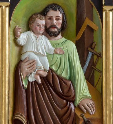 Zdjęcie nr 1: Drewniana płaskorzeźba św. Józefa z Dzieciątkiem w kształcie stojącego prostokąta, zamkniętego łukiem półkolistym, umieszczona w architektonicznej oprawie. Święty Józef ukazany w centrum kompozycji z Dzieciątkiem Jezus na prawym ręku, lewą wspierający się na blacie stołu stolarskiego. Twarz o ostro zaznaczonych rysach, z długim i prostym nosem, małymi ustami i oczami, okolona krótką brodą, policzki zarumienione. Włosy ciemnobrązowe, średniej długości, opadające kosmykami na czoło i zasłaniające uszy. Święty ubrany jest w zieloną suknię z długimi rękawami oraz brązowy płaszcz zarzucony na prawe przedramię i zasłaniający nogi, na stopach ma sandały. Dzieciątko zwrócone trzy czwarte w lewo, ukazane w pozycji siedzącej ze skrzyżowanymi nóżkami, z szeroko rozłożonymi na boki rączkami. Twarz okrągła o dziecięcych rysach, niebieskich oczach i silnie zarumienionych policzkach. Włosy krótkie i jasne. Ubrany jest w białą sukienkę z długimi rękawami, obwiedzioną na brzegach złotą lamówką. Draperia szat ułożona w kanciate, równolegle biegnące fałdy. Po prawej stronie obrazu stół stolarski z imadłem oraz narzędziami stolarskimi: piłą i kątownicą. Całość ukazana we wnętrzu architektonicznym z drewnianą podłogą, zielonym tłem i drewnianym słupem po prawej stronie. Rama architektoniczna, od dołu ujęta półwałkiem, flankowana kolumienkami podtrzymującymi trójkątny przyczółek. Struktura ramy malowana na kolor ciemnobrązowy, polichromia w odsłoniętych partiach ciała płaskorzeźby naturalistyczna, detale złocone.
