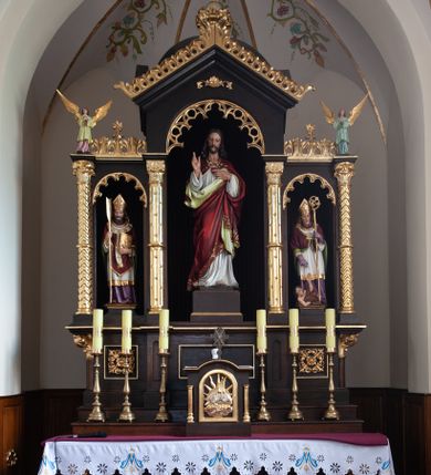 Zdjęcie nr 1: Nastawa ołtarzowa architektoniczna, trójosiowa, jednokondygnacyjna, na wysokim cokole. Ołtarz drewniany, prostopadłościenny; mensa drewniana. Antepedium w formie leżącego prostokąta ujęte w narożach kolumienkami, w polu trzy prostokątne płyciny, w środkowej monogram IHS na czerwonym tle otoczony promienistą glorią. Tabernakulum w formie prostopadłościennej skrzynki, ustawione na wysokim cokole, flankowane dwiem kolumienkami, zamknięte od góry gzymsem w kształcie łuku nadwieszonego. Drzwiczki w kształcie stojącego prostokąta, zamkniętego łukiem półkolistym, w polu płaskorzeźbione przedstawienie baranka paschalnego. Cokół nastawy trójosiowy, oś środkowa wydzielona pilastrami, na skrajach osi bocznych ćwierćkolumny z kapitelami w formie liści akantu. W polach osi bocznych kwadratowe płyciny wypełnione rozetami, w osi środkowej płycina w kształcie leżącego prostokąta. Cokół podtrzymuje nastawę, flankowaną dwiema parami kolumn, z których środkowe podtrzymują kopułę z przyczółkiem, a kolumny boczne figury aniołów. We wnęce środkowej figura Najświętszego Serca Jezusa, we wnęce lewej św. Wojciech, a po prawej św. Stanisław. Kolumny ustawione na bazach, przedzielone w połowie opaską, o trzonach zdobionych geometrycznymi motywami i kapitelach dekorowanych liśćmi akantu. U szczytu przyczółka duży kwiaton z zatkniętym krzyżem osadzonym na kuli. Gzymsy zdobione ażurową dekoracją roślinną. Łuki wnęk dekorowane arkadowaniami, w tym środkowa z maswerkami. Ponadto szczyt osi środkowej oraz gzyms nad osiami bocznymi wieńczy ornamentalny krenelaż. Struktura malowana w kolorze ciemnego brązu, ornamentyka i detale złocone. 

