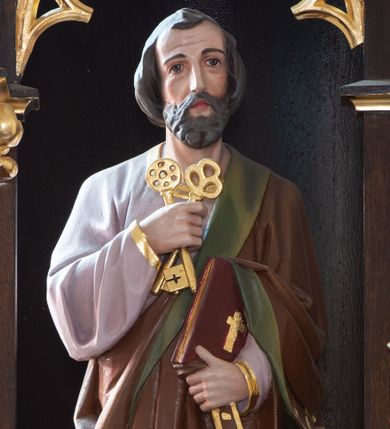 Zdjęcie nr 1: Rzeźba św. Piotra ustawiona na niskim prostopadłościennym cokole o ściętych narożach. Święty Piotr ukazany frontalnie, w delikatnym kontrapoście, z lewą nogą ugiętą w kolanie. W prawej dłoni trzyma dwa skrzyżowane klucze, a w lewej księgę. Twarz podłużna ze zmarszczonym czołem, z długim i szerokim nosem, dużymi oczami i małymi ustami, okolona średniej długości brodą. Włosy ciemnobrązowe, gęste i zasłaniające uszy. Ubrany jest w długą fioletową suknię z długimi rękawami oraz brązowy płaszcz z zieloną podszewką, zdobiony na brzegach dekoracyjnym pasem utworzonym z ukośnych kresek. Księga w czerwonej obwolucie, zdobiona złotym, łacińskim krzyżykiem. Polichromia w odsłoniętych partiach ciała naturalistyczna, detale złocone.