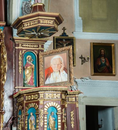 Zdjęcie nr 2: Ambona drewniana, przyścienna, dostępna jednobiegowymi schodkami od strony prezbiterium. Kosz na planie ośmioboku, na cokole dekorowanym złoconym ornamentem małżowinowym; artykułowany pilastrami wspierającymi fryz wypełniony złoconym ornamentem małżowinowym. Na trzonach pilastrów złocony ornament kwiatowy podwieszony na wstędze regencyjnej. W polach płyciny ujęte boniowanymi arkadkami mieszczącymi płaskorzeźbione, całopostaciowe przedstawienia ewangelistów na niebieskim tle. Całość zwieńczona profilowanym gzymsem. Zaplecek z półkolistą niszą mieszczącą płaskorzeźbione przedstawienie Chrystusa Dobrego Pasterza, ujętą pilastrami dźwigającymi niepełne belkowanie. Na trzonach podwieszone złocone zwisy kwiatowe przewiązane karbowaną wstęgą. Baldachim ośmioboczny z silnie występującym, profilowanym gzymsem. W podniebiu płaskorzeźbione przedstawienie gołębicy Ducha Świętego. W zwieńczeniu, na ośmiobocznym postumencie wolnostojąca, pełnoplastyczna rzeźba anioła grającego na trąbie. Figury ewangelistów ukazane frontalnie, w postawie stojącej, w dłoniach z których jedna jest złożona na piersi, trzymają pióra oraz zwoje lub księgi. Mają twarze podłużne, okolone brodami i średniej długości włosami. W pierwszej arkadzie od lewej strony ukazany jest św. Mateusz z małą postacią człowieka u nóg. Święty ubrany jest w brązową suknię oraz złoty płaszcz; ma siwe włosy. W kolejnej arkadzie św. Marek, a za nim lew. Święty ubrany jest w fioletową suknię i złocony płaszcz. W następnej arkadzie św. Łukasz, obok którego wyłania się wół. Ubrany jest w niebieską suknię i złocony płaszcz; ma siwe włosy i brodę. W ostatniej arkadzie ukazano św. Jana z orłem u stóp, ubranego w zieloną suknię i złocony płaszcz, bez zarostu, z długimi i jasnymi włosami. Chrystus ukazany frontalnie z barankiem założonym na barku, przytrzymujący go lewą ręką, z kijem pasterskim w prawej dłoni i drugim barankiem u stóp. Twarz podłużna o głęboko osadzonych, dużych oczach, okolona krótką brodą i długimi włosami. U szczytu anioł grający na trąbie. Ubrany jest w jasnofioletową suknię oraz krótką złoconą tunikę. Struktura malowana na ciemny brąz, partie ornamentu złocone.