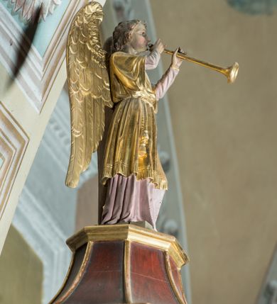 Zdjęcie nr 8: Ambona drewniana, przyścienna, dostępna jednobiegowymi schodkami od strony prezbiterium. Kosz na planie ośmioboku, na cokole dekorowanym złoconym ornamentem małżowinowym; artykułowany pilastrami wspierającymi fryz wypełniony złoconym ornamentem małżowinowym. Na trzonach pilastrów złocony ornament kwiatowy podwieszony na wstędze regencyjnej. W polach płyciny ujęte boniowanymi arkadkami mieszczącymi płaskorzeźbione, całopostaciowe przedstawienia ewangelistów na niebieskim tle. Całość zwieńczona profilowanym gzymsem. Zaplecek z półkolistą niszą mieszczącą płaskorzeźbione przedstawienie Chrystusa Dobrego Pasterza, ujętą pilastrami dźwigającymi niepełne belkowanie. Na trzonach podwieszone złocone zwisy kwiatowe przewiązane karbowaną wstęgą. Baldachim ośmioboczny z silnie występującym, profilowanym gzymsem. W podniebiu płaskorzeźbione przedstawienie gołębicy Ducha Świętego. W zwieńczeniu, na ośmiobocznym postumencie wolnostojąca, pełnoplastyczna rzeźba anioła grającego na trąbie. Figury ewangelistów ukazane frontalnie, w postawie stojącej, w dłoniach z których jedna jest złożona na piersi, trzymają pióra oraz zwoje lub księgi. Mają twarze podłużne, okolone brodami i średniej długości włosami. W pierwszej arkadzie od lewej strony ukazany jest św. Mateusz z małą postacią człowieka u nóg. Święty ubrany jest w brązową suknię oraz złoty płaszcz; ma siwe włosy. W kolejnej arkadzie św. Marek, a za nim lew. Święty ubrany jest w fioletową suknię i złocony płaszcz. W następnej arkadzie św. Łukasz, obok którego wyłania się wół. Ubrany jest w niebieską suknię i złocony płaszcz; ma siwe włosy i brodę. W ostatniej arkadzie ukazano św. Jana z orłem u stóp, ubranego w zieloną suknię i złocony płaszcz, bez zarostu, z długimi i jasnymi włosami. Chrystus ukazany frontalnie z barankiem założonym na barku, przytrzymujący go lewą ręką, z kijem pasterskim w prawej dłoni i drugim barankiem u stóp. Twarz podłużna o głęboko osadzonych, dużych oczach, okolona krótką brodą i długimi włosami. U szczytu anioł grający na trąbie. Ubrany jest w jasnofioletową suknię oraz krótką złoconą tunikę. Struktura malowana na ciemny brąz, partie ornamentu złocone.