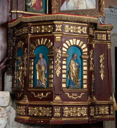 Zdjęcie nr 1: Ambona drewniana, przyścienna, dostępna jednobiegowymi schodkami od strony prezbiterium. Kosz na planie ośmioboku, na cokole dekorowanym złoconym ornamentem małżowinowym; artykułowany pilastrami wspierającymi fryz wypełniony złoconym ornamentem małżowinowym. Na trzonach pilastrów złocony ornament kwiatowy podwieszony na wstędze regencyjnej. W polach płyciny ujęte boniowanymi arkadkami mieszczącymi płaskorzeźbione, całopostaciowe przedstawienia ewangelistów na niebieskim tle. Całość zwieńczona profilowanym gzymsem. Zaplecek z półkolistą niszą mieszczącą płaskorzeźbione przedstawienie Chrystusa Dobrego Pasterza, ujętą pilastrami dźwigającymi niepełne belkowanie. Na trzonach podwieszone złocone zwisy kwiatowe przewiązane karbowaną wstęgą. Baldachim ośmioboczny z silnie występującym, profilowanym gzymsem. W podniebiu płaskorzeźbione przedstawienie gołębicy Ducha Świętego. W zwieńczeniu, na ośmiobocznym postumencie wolnostojąca, pełnoplastyczna rzeźba anioła grającego na trąbie. Figury ewangelistów ukazane frontalnie, w postawie stojącej, w dłoniach z których jedna jest złożona na piersi, trzymają pióra oraz zwoje lub księgi. Mają twarze podłużne, okolone brodami i średniej długości włosami. W pierwszej arkadzie od lewej strony ukazany jest św. Mateusz z małą postacią człowieka u nóg. Święty ubrany jest w brązową suknię oraz złoty płaszcz; ma siwe włosy. W kolejnej arkadzie św. Marek, a za nim lew. Święty ubrany jest w fioletową suknię i złocony płaszcz. W następnej arkadzie św. Łukasz, obok którego wyłania się wół. Ubrany jest w niebieską suknię i złocony płaszcz; ma siwe włosy i brodę. W ostatniej arkadzie ukazano św. Jana z orłem u stóp, ubranego w zieloną suknię i złocony płaszcz, bez zarostu, z długimi i jasnymi włosami. Chrystus ukazany frontalnie z barankiem założonym na barku, przytrzymujący go lewą ręką, z kijem pasterskim w prawej dłoni i drugim barankiem u stóp. Twarz podłużna o głęboko osadzonych, dużych oczach, okolona krótką brodą i długimi włosami. U szczytu anioł grający na trąbie. Ubrany jest w jasnofioletową suknię oraz krótką złoconą tunikę. Struktura malowana na ciemny brąz, partie ornamentu złocone.