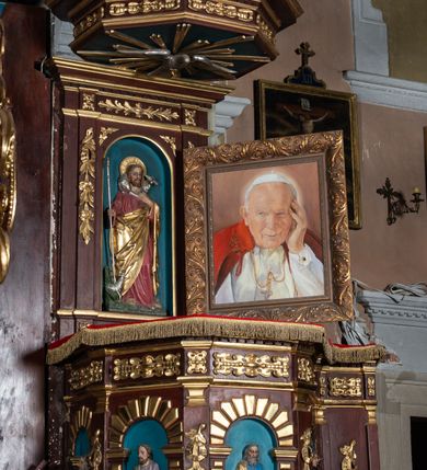 Zdjęcie nr 3: Ambona drewniana, przyścienna, dostępna jednobiegowymi schodkami od strony prezbiterium. Kosz na planie ośmioboku, na cokole dekorowanym złoconym ornamentem małżowinowym; artykułowany pilastrami wspierającymi fryz wypełniony złoconym ornamentem małżowinowym. Na trzonach pilastrów złocony ornament kwiatowy podwieszony na wstędze regencyjnej. W polach płyciny ujęte boniowanymi arkadkami mieszczącymi płaskorzeźbione, całopostaciowe przedstawienia ewangelistów na niebieskim tle. Całość zwieńczona profilowanym gzymsem. Zaplecek z półkolistą niszą mieszczącą płaskorzeźbione przedstawienie Chrystusa Dobrego Pasterza, ujętą pilastrami dźwigającymi niepełne belkowanie. Na trzonach podwieszone złocone zwisy kwiatowe przewiązane karbowaną wstęgą. Baldachim ośmioboczny z silnie występującym, profilowanym gzymsem. W podniebiu płaskorzeźbione przedstawienie gołębicy Ducha Świętego. W zwieńczeniu, na ośmiobocznym postumencie wolnostojąca, pełnoplastyczna rzeźba anioła grającego na trąbie. Figury ewangelistów ukazane frontalnie, w postawie stojącej, w dłoniach z których jedna jest złożona na piersi, trzymają pióra oraz zwoje lub księgi. Mają twarze podłużne, okolone brodami i średniej długości włosami. W pierwszej arkadzie od lewej strony ukazany jest św. Mateusz z małą postacią człowieka u nóg. Święty ubrany jest w brązową suknię oraz złoty płaszcz; ma siwe włosy. W kolejnej arkadzie św. Marek, a za nim lew. Święty ubrany jest w fioletową suknię i złocony płaszcz. W następnej arkadzie św. Łukasz, obok którego wyłania się wół. Ubrany jest w niebieską suknię i złocony płaszcz; ma siwe włosy i brodę. W ostatniej arkadzie ukazano św. Jana z orłem u stóp, ubranego w zieloną suknię i złocony płaszcz, bez zarostu, z długimi i jasnymi włosami. Chrystus ukazany frontalnie z barankiem założonym na barku, przytrzymujący go lewą ręką, z kijem pasterskim w prawej dłoni i drugim barankiem u stóp. Twarz podłużna o głęboko osadzonych, dużych oczach, okolona krótką brodą i długimi włosami. U szczytu anioł grający na trąbie. Ubrany jest w jasnofioletową suknię oraz krótką złoconą tunikę. Struktura malowana na ciemny brąz, partie ornamentu złocone.