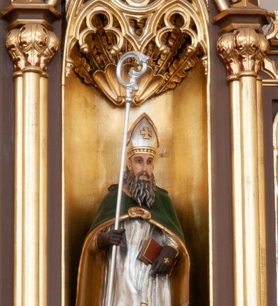 Zdjęcie nr 3: Ołtarz przyścienny, architektoniczny, pięcioosiowy, jednokondygnacyjny ze zwieńczeniem. Antepedium dekorowane płyciną w profilowanej ramie z umieszczonym centralnie czteroliściem oraz krzyżem. Tabernakulum metalowe, jednodrzwiczkowe z umieszczonym centralnie kielichem z hostią na tle kłosów oraz winorośli. Po bokach umieszczone litery „A” i „Ω” na tle krzyża każda. Nastawa posadowiona na wysokim cokole z płycinami wypełnionymi maswerkiem. Osie w układzie schodkowym z osią środkową wyższą i szerszą od bocznych, wydzielone wiązkami służek na postumentach w formie skręconych trzonów, z kapitelami dekorowanymi motywem ostroliścia. Służki zwieńczone pinaklami zakończonymi kwiatonami, w osi środkowej ażurowa wieżyczka w formie zbliżonej do pinakla zakończonego kwiatonem i krzyżem, tworząca baldachim nad figurą Dzieciątka Jezus. Osie środkowe zwieńczone trójkątnymi wimpergami wypełnionymi dekoracją maswerkową. W polu ołtarza obraz Trójcy Świętej w złoconej, profilowanej ramie w kształcie stojącego prostokąta zamkniętego łukiem ostrym z maswerkiem. Poniżej płycina z płaskorzeźbionym krzyżem na tle kłosów. W osiach drugiej i czwartej złocone nisze mieszczące figury św. Stanisława i Wojciecha. Nisze zamknięte od góry muszlami palmetowymi. Pod niszami płyciny wypełnione maswerkiem z fantazyjnym motywem geometrycznym. Osie skrajne wypełnione zdwojonymi blendami z rozetami pośrodku. Struktura ołtarza malowana na brązowo z elementami czerwieni, detal architektoniczny złocony. Figury w pełni polichromowane.