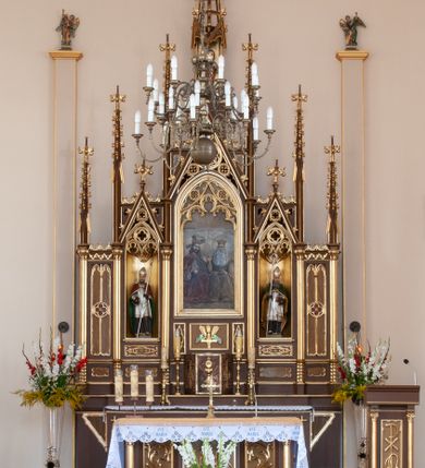 Zdjęcie nr 1: Ołtarz przyścienny, architektoniczny, pięcioosiowy, jednokondygnacyjny ze zwieńczeniem. Antepedium dekorowane płyciną w profilowanej ramie z umieszczonym centralnie czteroliściem oraz krzyżem. Tabernakulum metalowe, jednodrzwiczkowe z umieszczonym centralnie kielichem z hostią na tle kłosów oraz winorośli. Po bokach umieszczone litery „A” i „Ω” na tle krzyża każda. Nastawa posadowiona na wysokim cokole z płycinami wypełnionymi maswerkiem. Osie w układzie schodkowym z osią środkową wyższą i szerszą od bocznych, wydzielone wiązkami służek na postumentach w formie skręconych trzonów, z kapitelami dekorowanymi motywem ostroliścia. Służki zwieńczone pinaklami zakończonymi kwiatonami, w osi środkowej ażurowa wieżyczka w formie zbliżonej do pinakla zakończonego kwiatonem i krzyżem, tworząca baldachim nad figurą Dzieciątka Jezus. Osie środkowe zwieńczone trójkątnymi wimpergami wypełnionymi dekoracją maswerkową. W polu ołtarza obraz Trójcy Świętej w złoconej, profilowanej ramie w kształcie stojącego prostokąta zamkniętego łukiem ostrym z maswerkiem. Poniżej płycina z płaskorzeźbionym krzyżem na tle kłosów. W osiach drugiej i czwartej złocone nisze mieszczące figury św. Stanisława i Wojciecha. Nisze zamknięte od góry muszlami palmetowymi. Pod niszami płyciny wypełnione maswerkiem z fantazyjnym motywem geometrycznym. Osie skrajne wypełnione zdwojonymi blendami z rozetami pośrodku. Struktura ołtarza malowana na brązowo z elementami czerwieni, detal architektoniczny złocony. Figury w pełni polichromowane.